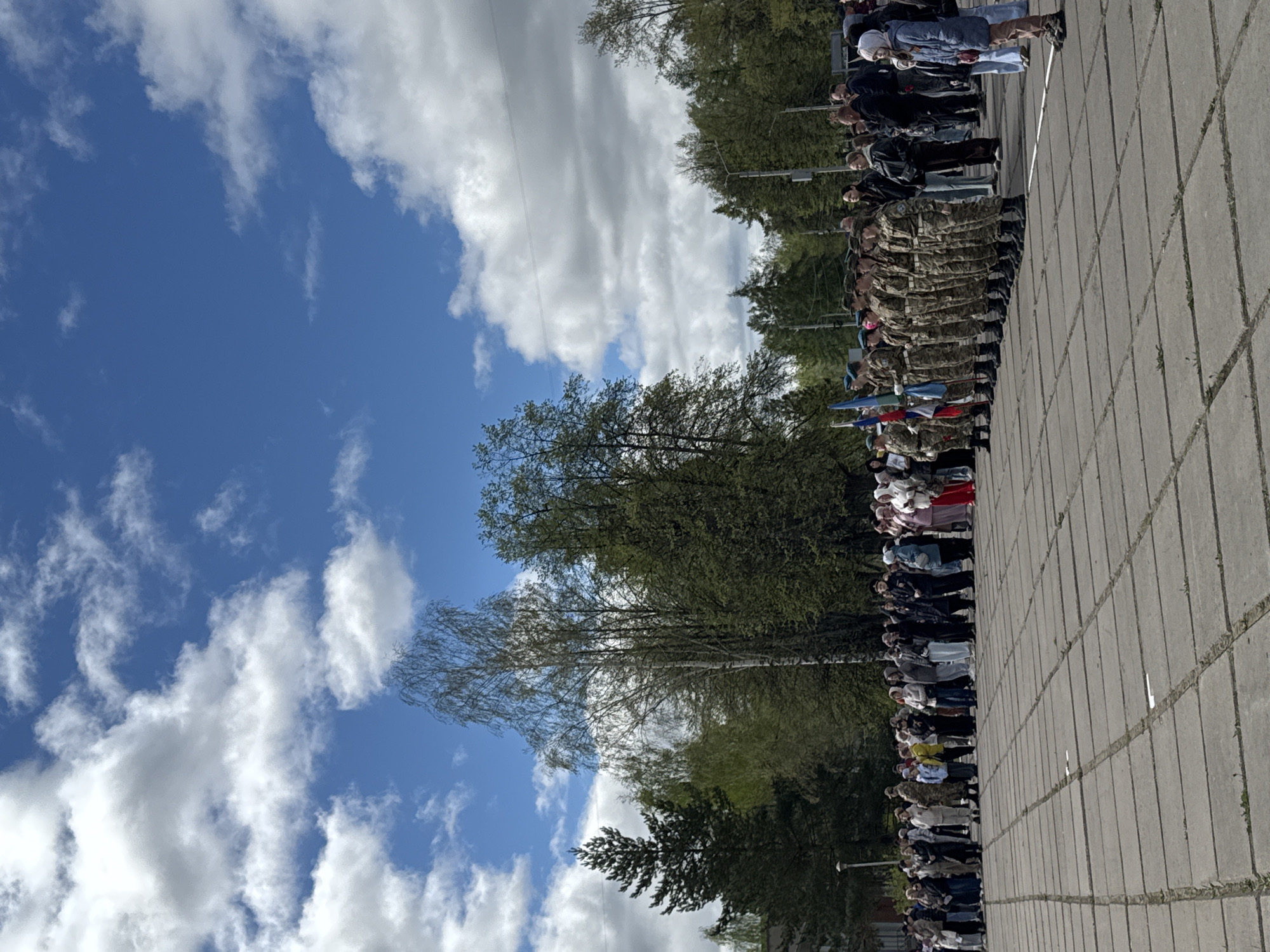 The image shows a military parade or commemoration event in what is likely the city of Chaikovsky, Russia. A line of uniformed military personnel stand at attention, holding flags. A crowd of civilians, likely spectators, is present, some wearing head coverings. The event takes place on a paved area with trees visible in the background and a blue sky with white clouds overhead. Overhead electrical wires are visible.