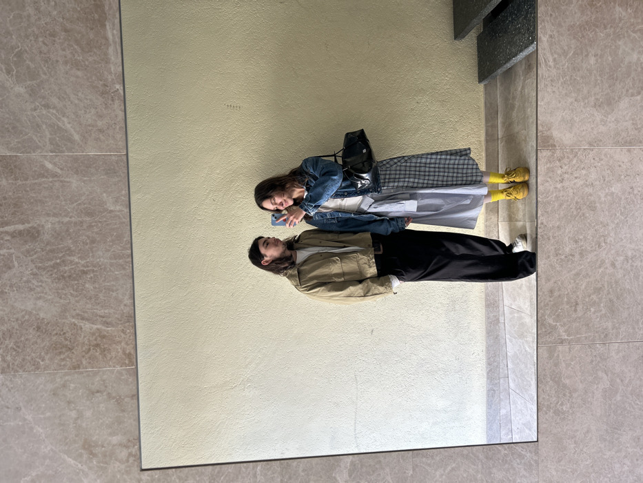 The image depicts a mirror selfie of two individuals taken in an indoor setting within Barcelona, Spain. The reflection shows a textured, light yellow wall as the background and a light grey tiled floor. To the far right of the reflection, dark, multi-level steps are partially visible. The mirror itself is bordered by light brown marble tiles at the top and bottom.
A male-presenting individual stands on the left, with long dark hair, wearing a light-colored long-sleeved shirt beneath a beige, collared, front-pocketed jacket, black wide-leg trousers, and white sneakers with white socks. He is oriented towards the individual on his right.
A female-presenting individual stands on the right, also with long dark hair. She holds a dark-colored smartphone with both hands, angled towards the mirror, indicating she is capturing the reflection. She wears a blue denim jacket over a white top, a long skirt featuring a light blue solid section on one side and a grey plaid pattern on the other. Her footwear consists of bright yellow socks and yellow sneakers. A black shoulder bag is suspended from her left shoulder. She is smiling and appears to be looking at the phone screen.