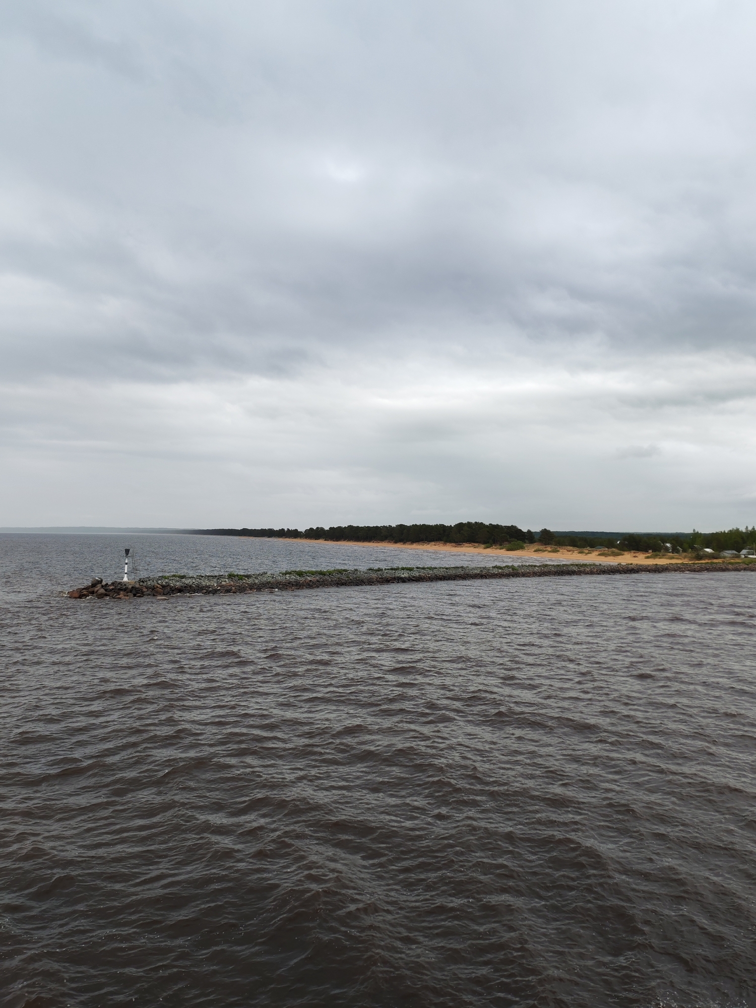 The image depicts a seascape under an overcast sky, possibly in the vicinity of Ozernoe Ust'e, Russia. In the foreground, dark, choppy water stretches towards a long, rocky breakwater that extends into the sea. The breakwater is topped with sparse green vegetation and a white and black navigational marker is at its end. Beyond the breakwater, a shoreline of sandy beach is backed by a dense band of coniferous trees. A few buildings are visible in the distance.