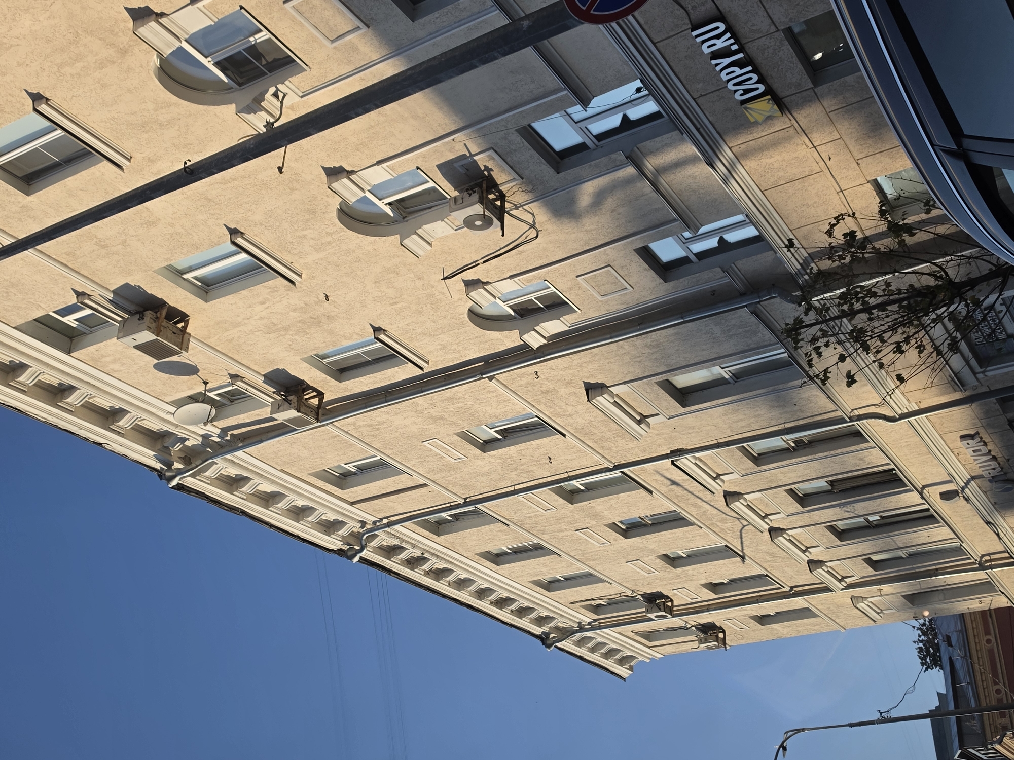 An angled, medium shot captures a section of a light tan, multi-story building on a sunny day. The building's facade is textured, with decorative white trim around the windows and along the top edge. Several air conditioning units are mounted on the exterior walls. To the right of the building, a dark grey utility pole runs vertically, casting a shadow. A partial view of a dark colored car is in the lower right foreground. A sign reading "COPY.RU" is visible on the building's lower level. The sky above is a clear, bright blue. The image is taken from a low angle looking upwards. Based on visual cues such as architecture and the presence of the "COPY.RU" sign, this location is likely in Nizhny Novgorod, Russia.