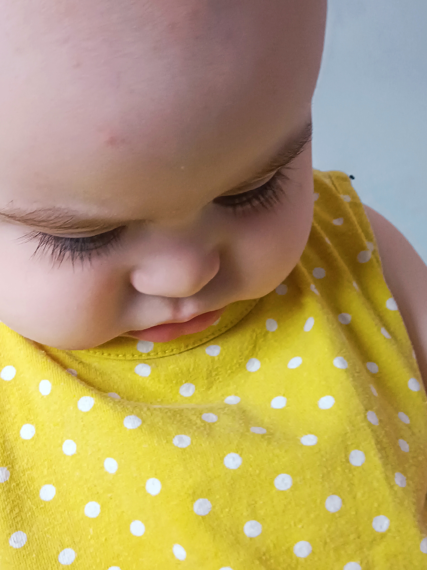 A close-up, eye-level shot focuses on the face of a young child, likely a toddler. The child is looking downwards, their head tilted to the right. The dominant colors are the warm tones of the child's skin and a bright yellow garment adorned with white polka dots. The child's features are softly lit, highlighting their closed eyes with long, dark eyelashes and a small, slightly parted mouth. A faint reddish mark is visible on the forehead. The background is a soft, pale blue, suggesting an indoor environment with plain walls. The image captures an intimate and tender moment, devoid of any specific urban or environmental markers that would indicate a location within Noviy, Russia.