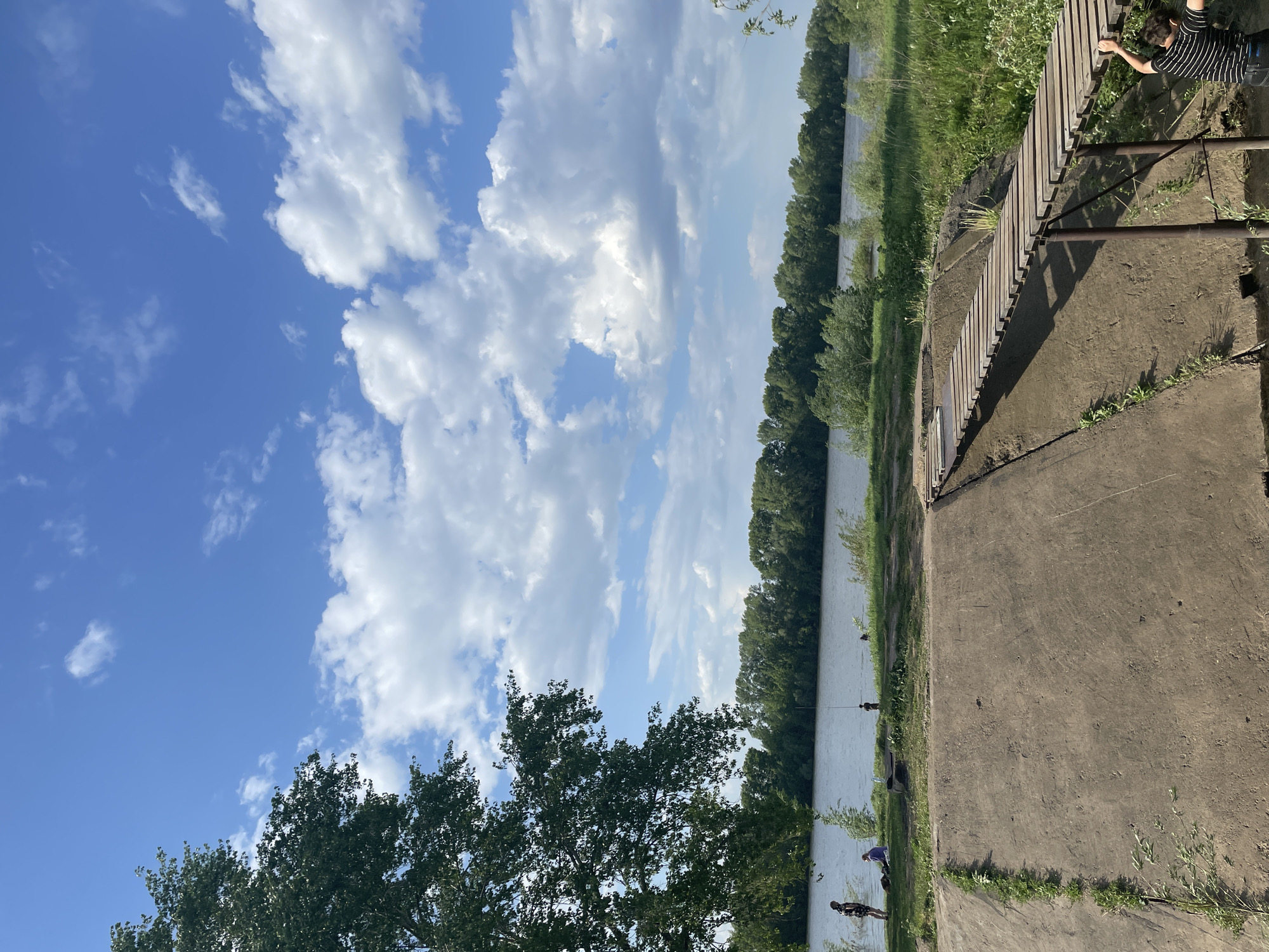 The image depicts an outdoor scene with a body of water, likely a river or wide canal, running along the middle. On the right side is a concrete embankment with a wooden railing. A person is leaning over the railing. On the left, the view opens to a sky filled with fluffy white clouds against a blue backdrop, with some trees at the bottom of the frame. Further down the riverbank, there are some individuals. The location is in the city of Unknown, Unknown.