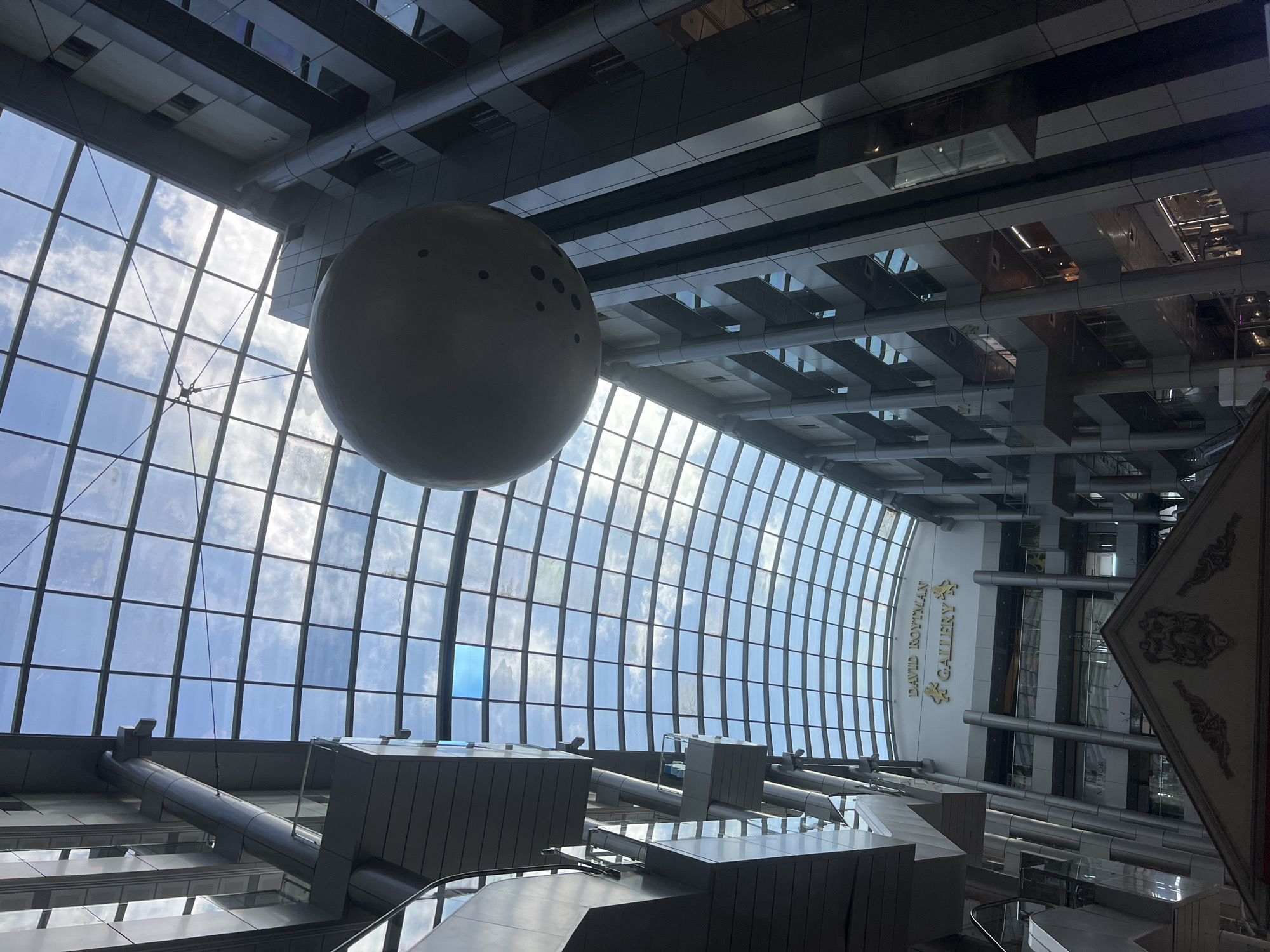 The image captures the interior of a modern atrium in Odesa, Ukraine, likely a shopping mall or gallery. A large, spherical sculpture, resembling a planet with craters, hangs suspended from the glass ceiling by wires. The atrium is multi-storied, with a predominantly grey and metallic facade. A large, arched glass ceiling allows natural light to filter through, illuminating the space and revealing a partly cloudy sky. A sign reading "DAVID ROYTMAN GALLERY" is visible on the upper level, indicating the presence of an art gallery. Escalators and walkways connect the different floors. The overall impression is one of contemporary architecture and indoor public space.