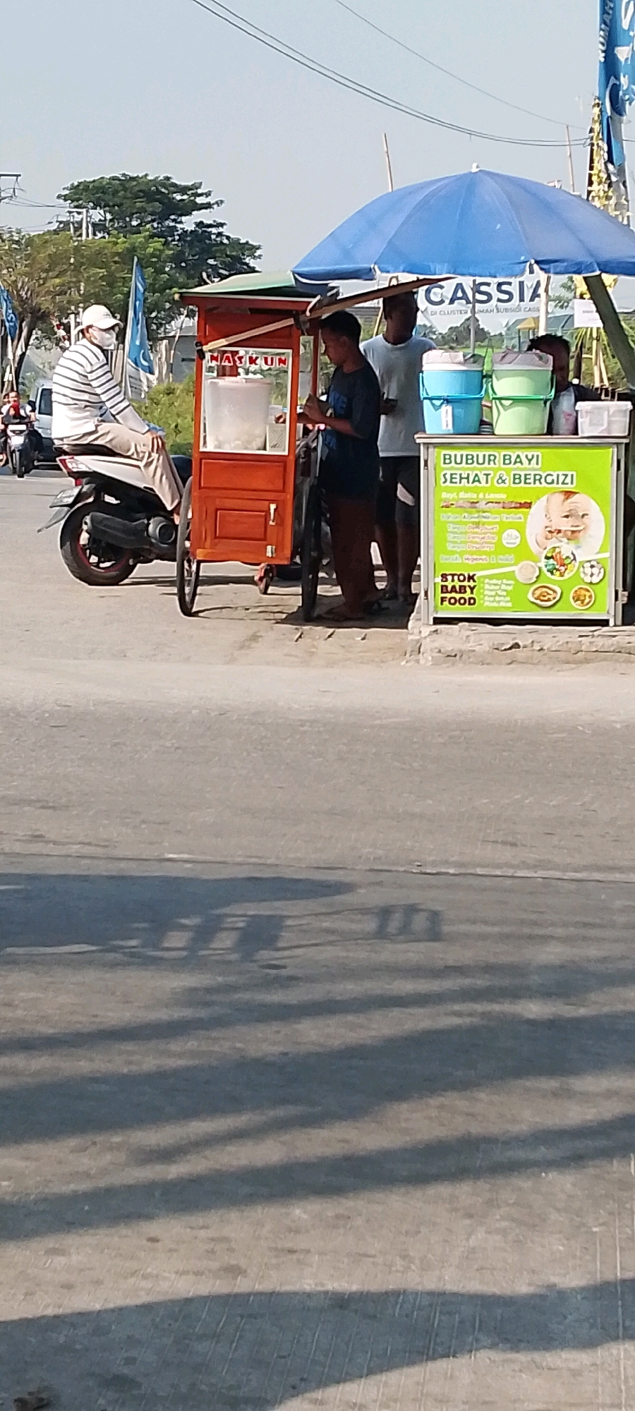 In Sriamur, Indonesia, a street scene unfolds with a food cart and several individuals. A person wearing a striped shirt and a white cap sits on a motorcycle, facing away from the viewer. To the right of the motorcycle, a red food cart is positioned. A young man in a dark shirt and brown patterned pants stands by the cart, appearing to be engaged in preparing or serving food. Next to him, another young man in a light blue t-shirt stands, and partially obscured behind him is a third individual.
In front of the cart, a green sign is displayed with Indonesian text and an illustration of a baby. The sign advertises "BUBUR BAYI SEHAT & BERGIZI" (Healthy & Nutritious Baby Porridge) and lists "STOK BABY FOOD." Two plastic buckets, one blue and one green, are stacked on top of each other near the food cart. A large blue umbrella is deployed above the food cart area, providing shade. In the background, trees and utility poles are visible, along with a flag with blue and white elements. The scene is lit by bright sunlight, casting distinct shadows on the asphalt road in the foreground. Several motorcycles are parked or moving in the background.