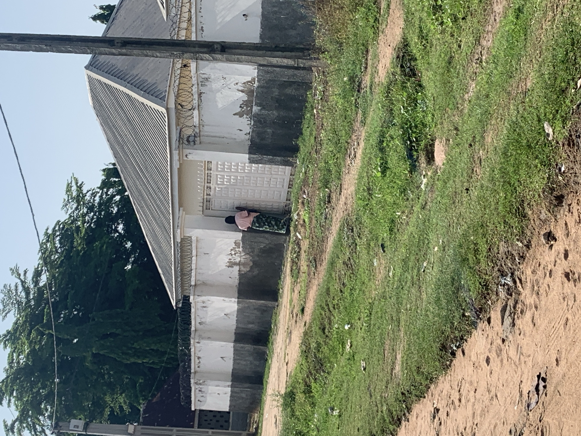The image captures a person, likely a woman, standing near a white gate in front of a building. She is wearing a pink shirt and a patterned skirt. The building appears to be partially aged, with peeling paint on its exterior walls and barbed wire along the top. A corrugated metal roof is visible on a section of the building. To the right of the building, there is a patch of overgrown green grass and a dirt path. The scene is bathed in natural light, suggesting it is daytime. The overall impression is of an outdoor setting, possibly in a less developed area, given the condition of the building and the presence of the grassy area. There is no visible text in the image.