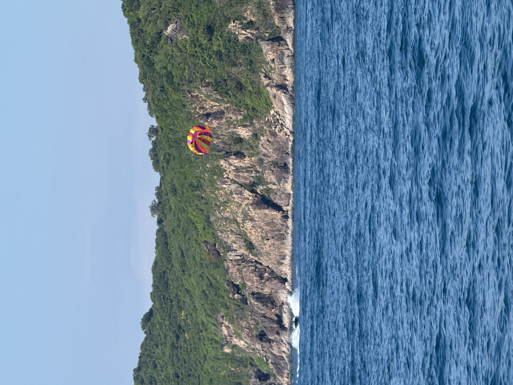 A parasailer, suspended from a colorful parachute, is visible in the sky above the ocean near a rocky, tree-covered island. A speedboat is creating a wake as it travels along the water. The scene takes place in the ocean off the coast of Bac Nha Trang Ward, Vietnam.