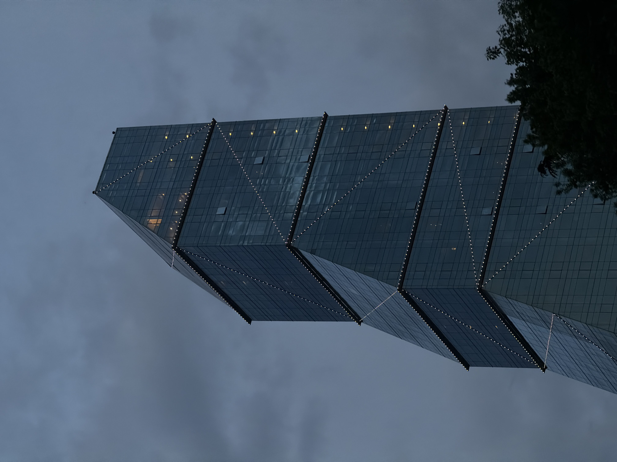 An angled close-up view shows the modern, glass-paneled Axis Towers building in Tbilisi, Georgia against a cloudy, twilight sky. The building's angular design features several distinct, protruding sections linked with lines of lights. There is a dark tree in the upper right corner of the frame. The scene is an urban nightscape.