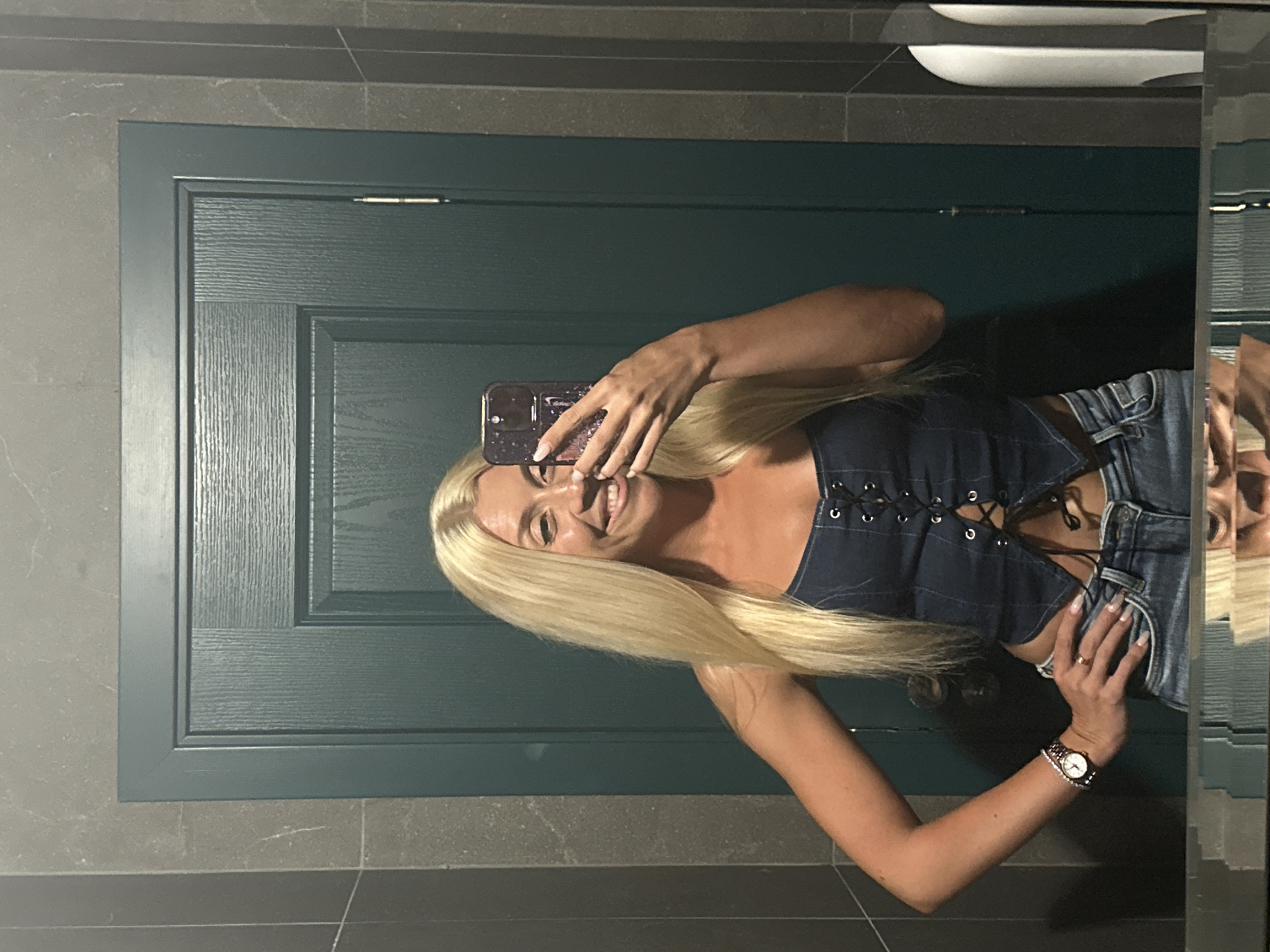 A young blonde woman takes a mirror selfie in a dimly lit, modern bathroom. She is wearing a blue denim corset-style top with black lace-up detailing and low-rise blue jeans. Her left arm is akimbo, and she is holding her phone in her right hand, angled to capture her reflection. She smiles and touches her face with her fingers, displaying a diamond-studded phone case. A watch adorns her left wrist. The bathroom features dark grey tiled walls and a dark teal-colored door. The reflection in the mirror shows the lower half of the woman and the bathroom counter. There is no indication of the city of Pazardzhik, Bulgaria, in the image.