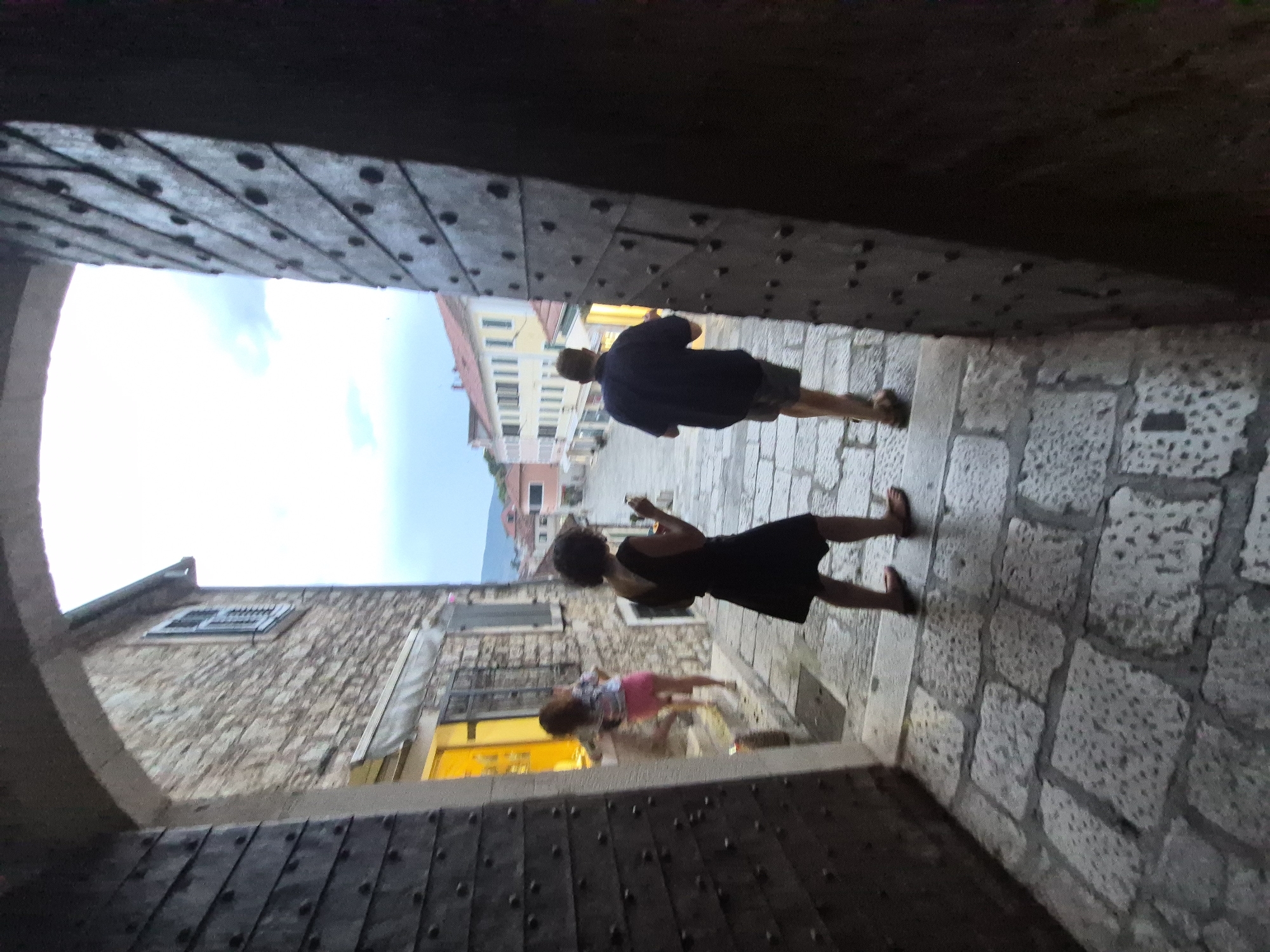 The image depicts three people walking through a stone archway, possibly an entrance to a historic town. The archway is made of heavy, dark wood with visible rivets. The ground is paved with cobblestones.
In the foreground, on the right, a woman in a black dress is walking away from the camera, holding a phone in her hand. To her left and slightly further ahead, a young child in a pink skirt and a patterned top is also walking away from the camera. Further ahead, a man in a dark shirt and shorts is walking away, also looking at his phone.
Beyond the archway, buildings with stone facades and tiled roofs are visible, suggesting an old European town setting. The sky is bright and appears to be daytime, possibly late afternoon given the angle of light and shadows. The overall scene suggests a casual stroll through a picturesque location. The location context provided is Herceg Novi, Montenegro. There is no visible text in the image.