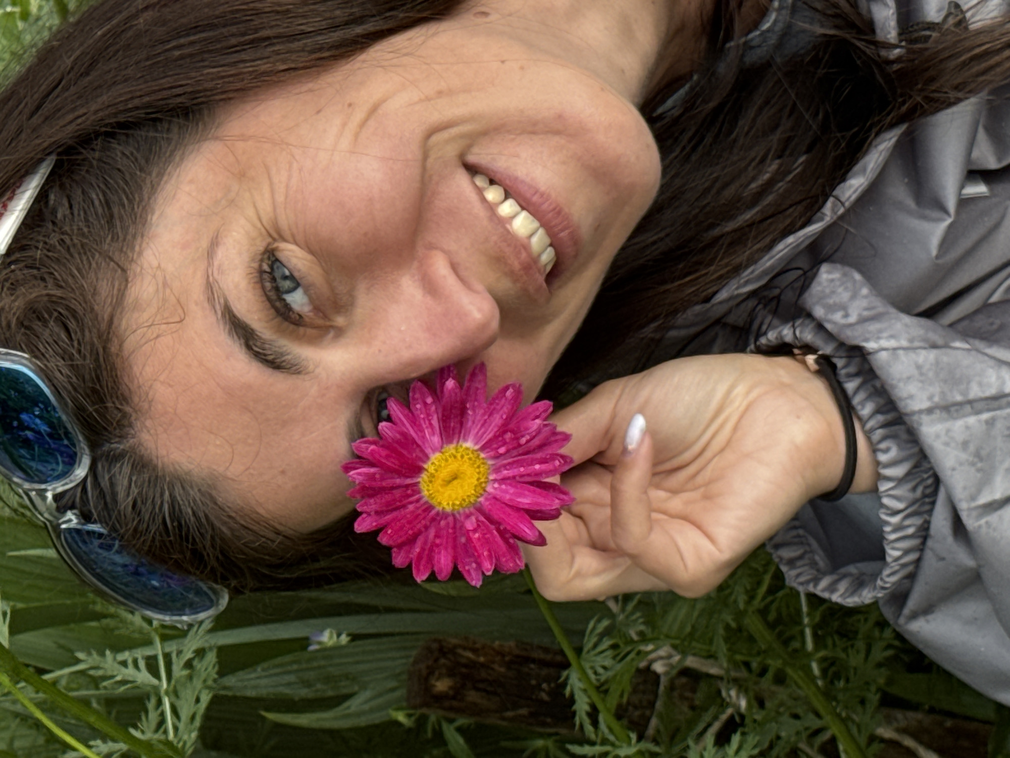 A woman with brown hair and blue eyes holds a bright pink daisy in front of her right eye. She is wearing sunglasses on her head and a grey jacket. The background consists of lush green foliage and a wooden post. There is no indication of the specific location within Vas'yata, Russia.