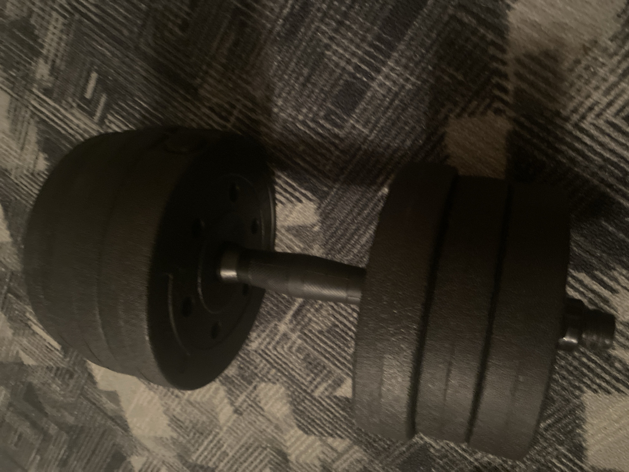 A black dumbbell is resting on a gray and white plaid blanket. The dumbbell has multiple weight plates stacked on either side of a central handle. The plates appear to be made of cast iron and have a textured surface. The handle is knurled for grip. The lighting in the image is dim, casting shadows on the blanket. The scene appears to be indoors, likely in a residential setting. There are no people or other objects visible in the image. The location in Chernogorsk, Russia is not discernible from the image.
