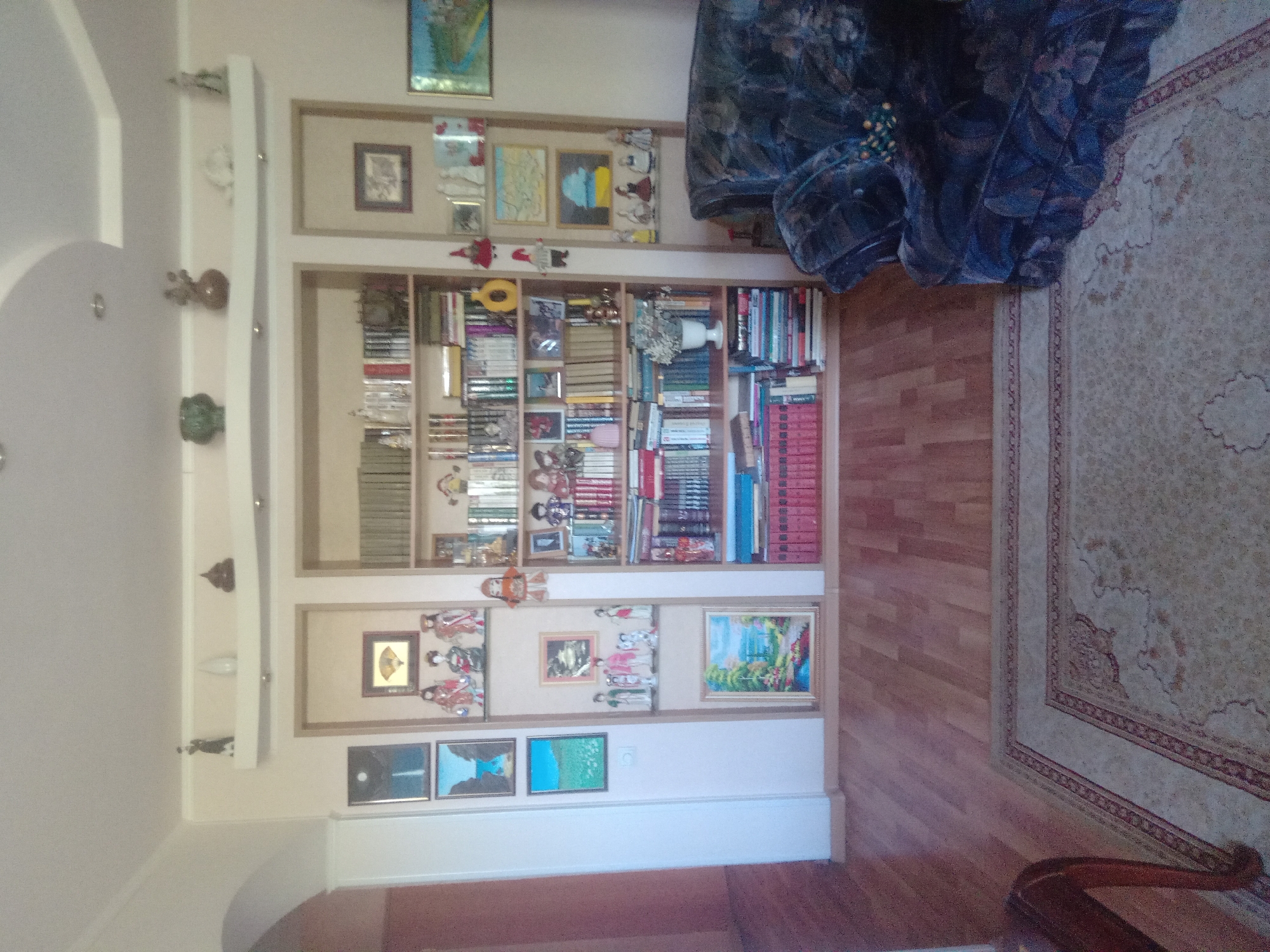 The image shows the interior of a room, likely a living room or study, in an apartment or house located in Oskemen, Kazakhstan. The room features a large built-in bookshelf filled with books, decorative items, and framed pictures. The bookshelf is the central focus, displaying various volumes, figurines, and artwork. To the left of the bookshelf, there is a white wall with several framed pictures arranged vertically. In the foreground, a patterned rug covers a portion of the wooden floor. To the right of the bookshelf, a dark-colored armchair is visible. The overall impression is of a well-decorated and organized living space.