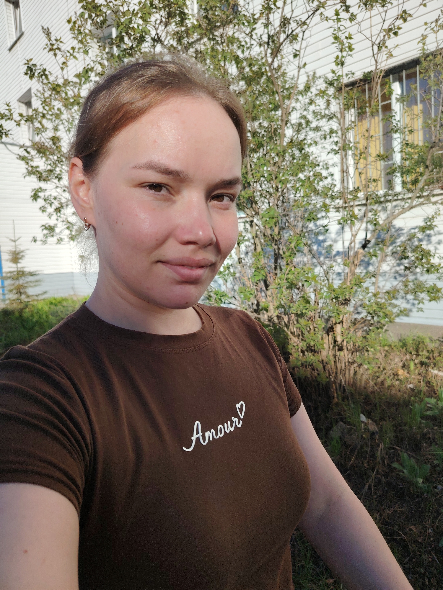 A fair-skinned young woman stands outdoors in front of a white, siding-covered building with windows. In the immediate background, a deciduous bush with small green leaves partially obscures the building. The woman is wearing a brown t-shirt with the word "Amour" printed in white script. She is looking at the camera with a slight smile. The environment appears to be a residential area. The image was taken in Chaikovsky, Russia.