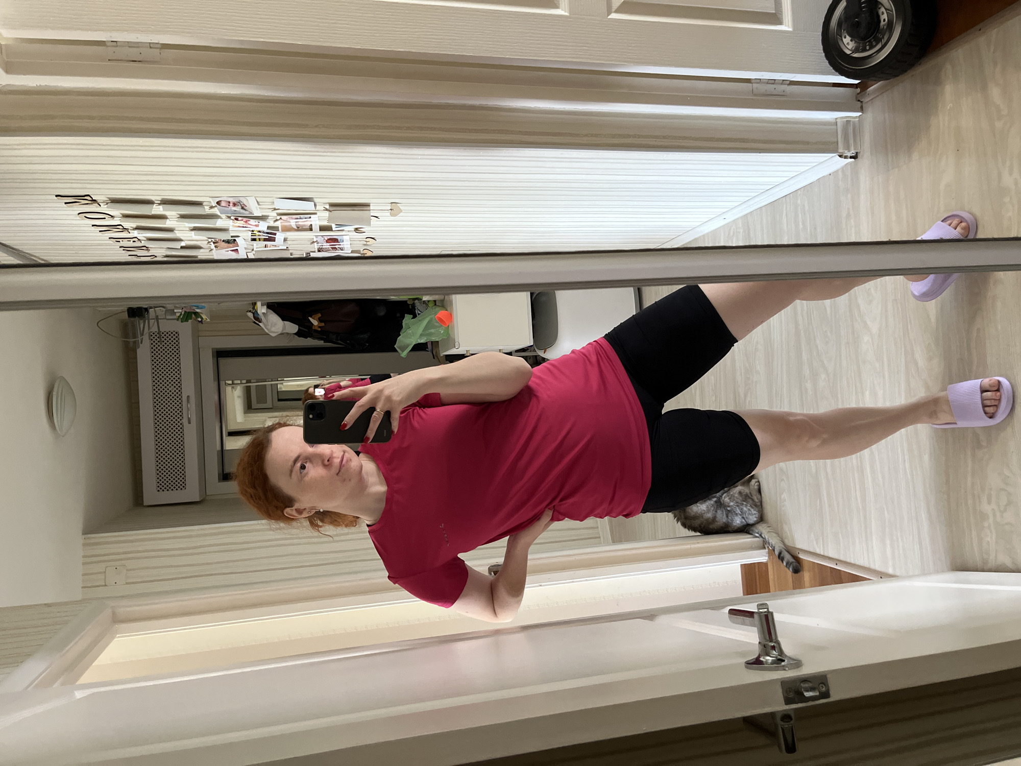 A woman with red hair, wearing a red t-shirt and black shorts, is taking a selfie in a mirror. She is holding a black smartphone in her right hand. She is standing in what appears to be a hallway. There is a cat visible near her legs. In the background, a cabinet with a scooter on top is visible. Another room is visible in the reflection, including a white cabinet and assorted items. Photos and writing are displayed on the top of a closet. The location is Chaikovsky, Russia.