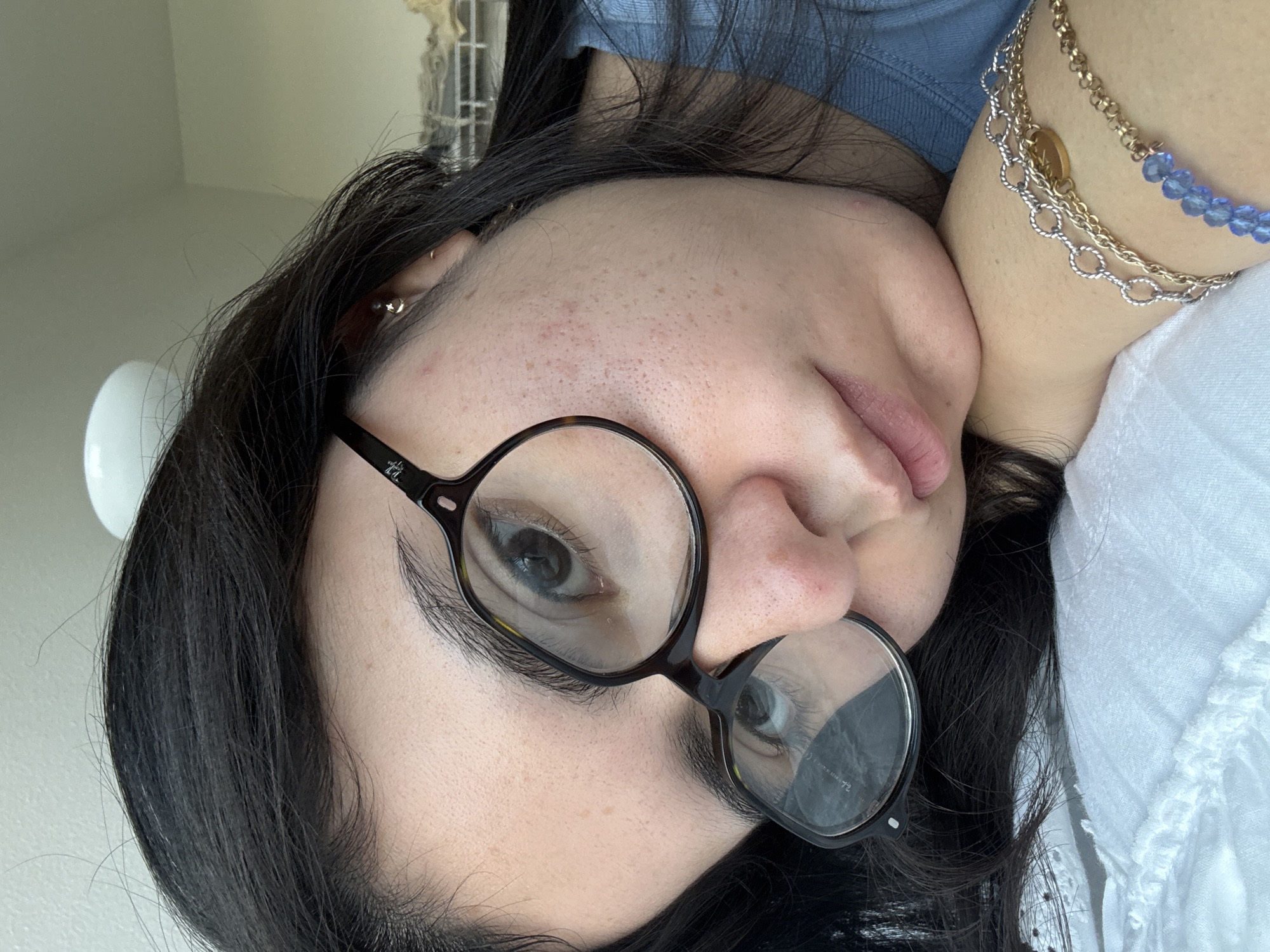 A close-up, upside-down portrait features a person with dark hair and fair skin, likely indoors in Austin, United States of America. The subject is wearing round, tortoise-shell eyeglasses, through which one eye with dark blue or grey iris and black eyeliner is clearly visible. The other eye is partially obscured by the glasses frame. The skin shows texture with visible pores and some minor blemishes on the forehead and cheek.
The person is resting their head on their hand or arm, conveying a relaxed posture. They wear multiple pieces of jewelry: small, shiny earlobe piercings (one appearing as a silver ball stud, another as a small gold hoop), and layered necklaces. The necklaces include a fine gold chain, a silver chain with a small, circular golden charm, and a beaded necklace with light blue, faceted stones.
The subject is wearing a light blue top, possibly ribbed, and a portion of a white garment with subtle texture is visible on the right side of the frame. The background is softly blurred, indicating an interior setting, possibly with ambient natural light. The brand "Ray-Ban" is faintly visible on the arm of the eyeglasses. No specific activity beyond resting is depicted.