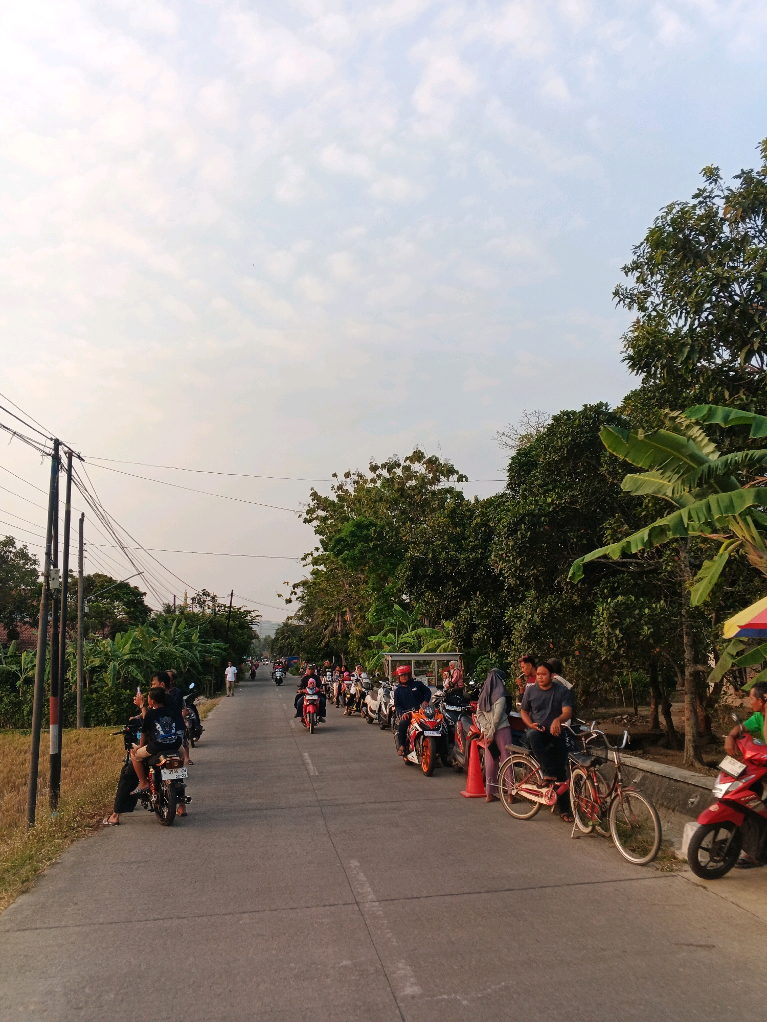 The image depicts a street scene in Kuripan, Indonesia, on a clear day with scattered clouds. A paved road runs through the center of the frame, lined with trees and vegetation on both sides. A group of people are gathered along the roadside, many of them on or around motorcycles.
On the left side of the road, several people are seated on motorcycles parked close to the edge. A utility pole with multiple wires is prominent in the foreground. The ground beside the road is covered in dry grass.
On the right side of the road, a longer line of motorcycles is parked. A red bicycle is positioned between two motorcycles. Several individuals are standing or sitting near these vehicles, some engaged in conversation. A colorful umbrella is visible further down the road on the right.
In the distance, more motorcycles are seen traveling along the road, and trees form a dense canopy on either side, with some glimpses of buildings and a mosque tower. The overall atmosphere suggests a casual gathering or a pause in travel. The lighting indicates it is likely late afternoon or early evening.