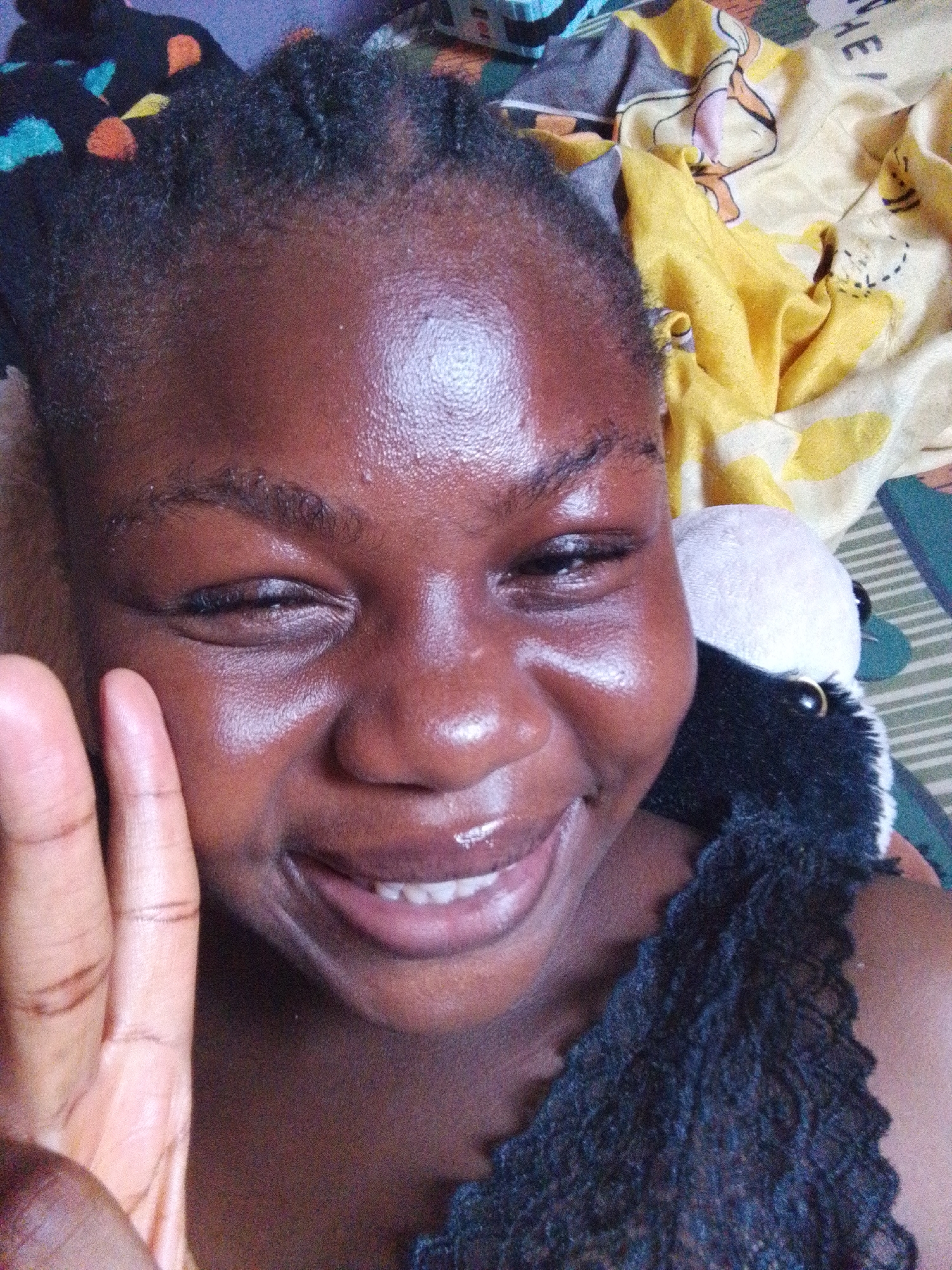A close-up selfie captures a young Black woman with dark brown skin and braided hair. Her skin appears shiny, possibly due to sweat or skincare products. She is smiling, revealing her teeth, and partially closing her eyes. Her right hand is raised with fingers spread. Adjacent to her face is a stuffed animal, seemingly a panda with black fur and a white head. Behind her are patterned bed sheets: a yellow sheet with cartoon characters and a striped sheet. The scene is indoors, likely a bedroom. No information is available regarding the location in the city of Nsukka, Nigeria.