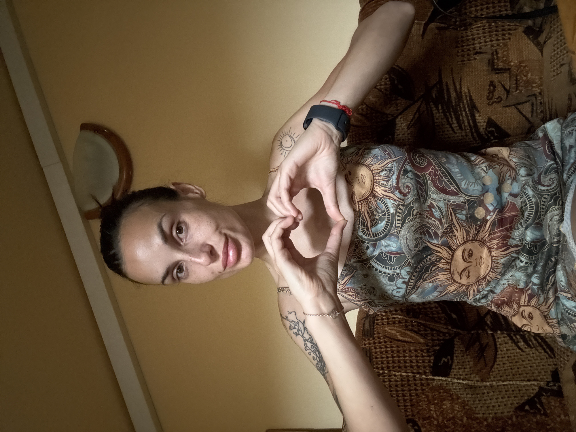 A woman with brown hair pulled back is seated on a brown couch, forming a heart shape with her hands. She is wearing a dress with a sun and paisley pattern. She has freckles and tattoos on her arms. The background is a yellowish wall with a light fixture. The image is taken indoors, likely in a residential setting in Pleven, Bulgaria.