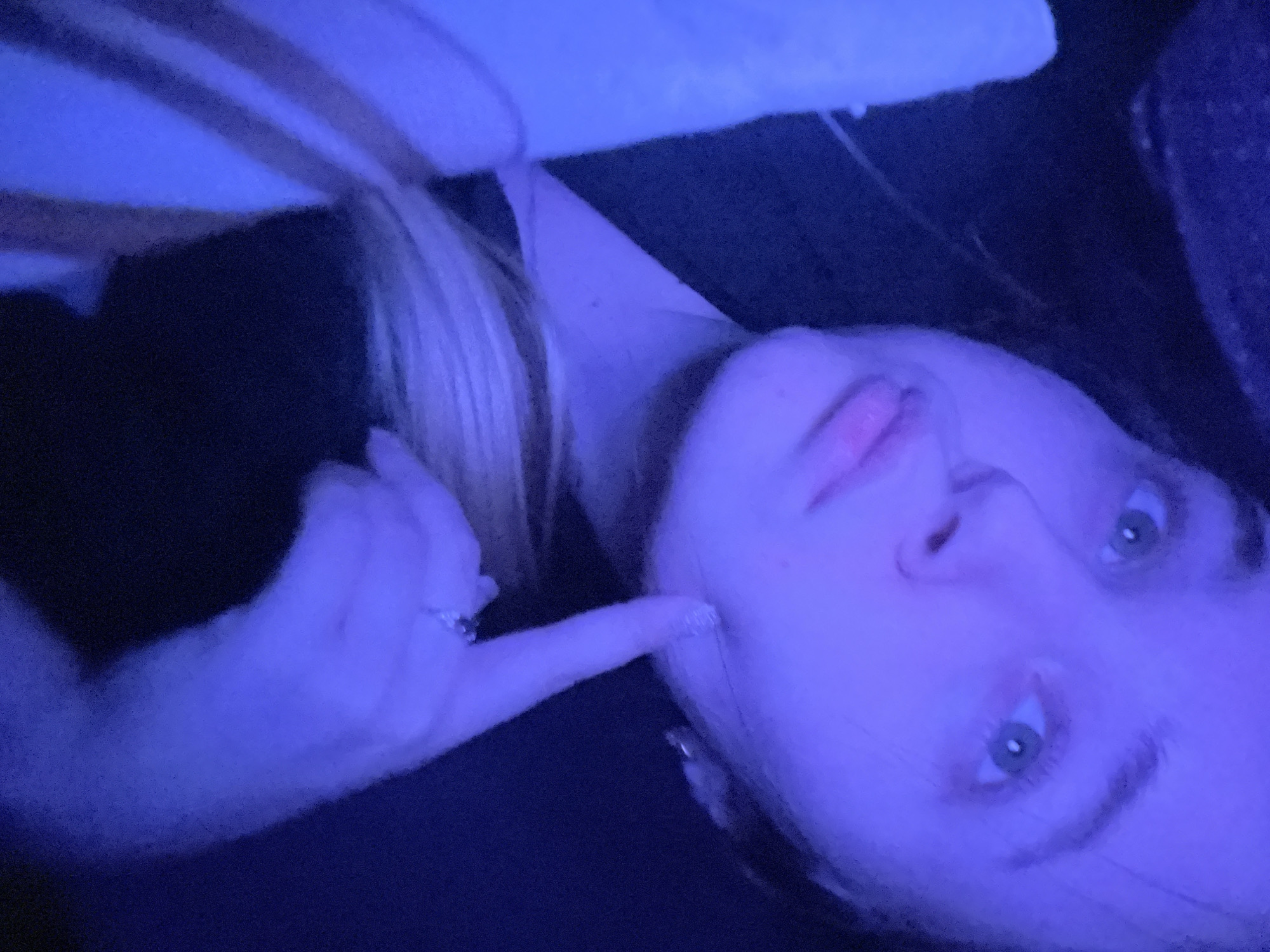 The image presents a close-up of a female subject under strong blue ambient lighting, resulting in a monochromatic blue cast over the scene. The subject's head and upper torso are visible, positioned at an upward angle, with her gaze directed towards the upper left. Her right index finger, adorned with a ring featuring multiple small stones, is lightly pressed against her right cheekbone. The subject has light blonde hair, visible over her right shoulder and partially framing her face, and light-colored eyes. She appears to be wearing dark clothing. In the lower right corner, a light-colored textile object, possibly a blanket with faint stripes, is present. The background is uniformly dark and indistinct, offering no specific environmental details. The image contains no visual information to confirm the specified location as Pushchino, Russia.