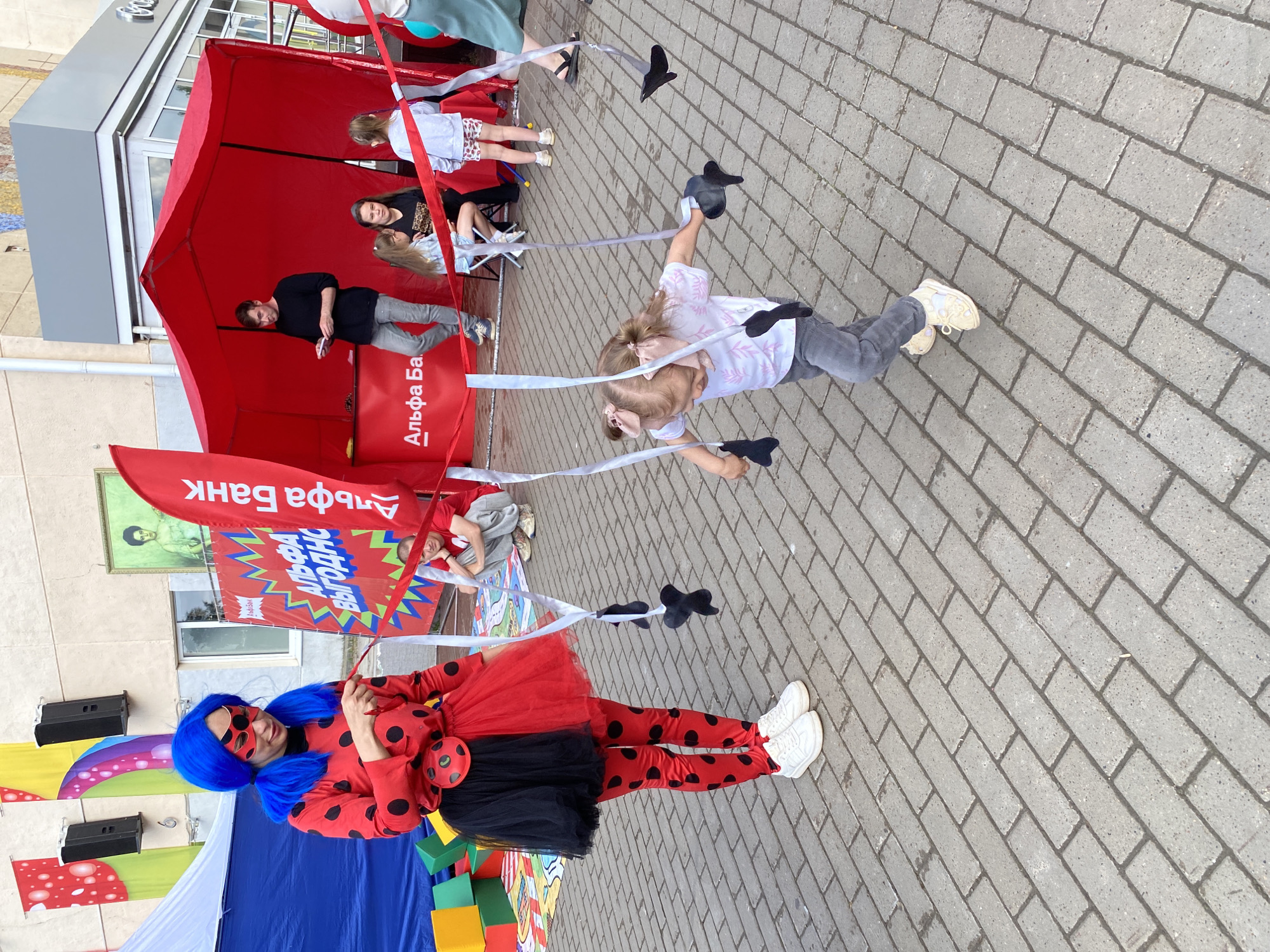 In an outdoor urban setting, likely a plaza or pedestrian area in Chaikovsky, Russia, a woman dressed as a ladybug character with a blue wig and red and black spotted costume, is holding a red ribbon. She is interacting with a young girl, who is running and holding onto several hanging ribbons that appear to be part of a game or activity. The girl is wearing a white t-shirt with a pink leaf pattern and gray pants. Behind them, a red tent with "Альфа Банк" (Alfa-Bank) branding is set up. Several other people are visible in the background, including a man in a black shirt and gray pants standing inside the tent and a man and woman seated on chairs. The ground is paved with gray brickwork. Colorful banners and speakers are mounted on a building in the background.