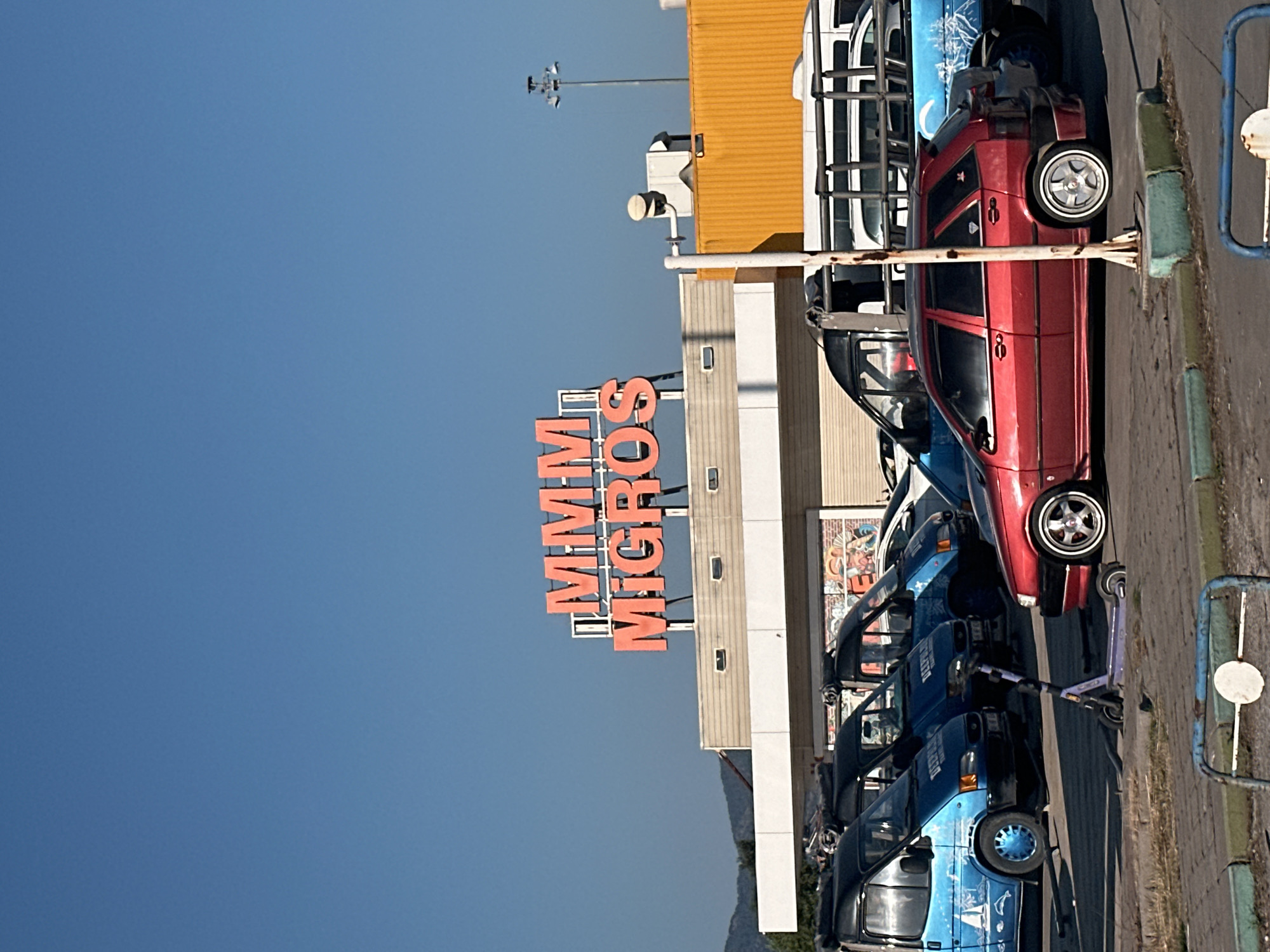 The image shows the "MMM Migros" sign in Fethiye, Turkey. Below the sign is the building of the Migros supermarket with a yellow adjacent building. Parked in front of the building are several blue vans, a red car, and a purple scooter. A blue metal barrier is visible at the far right of the image. The sky is a clear blue.
