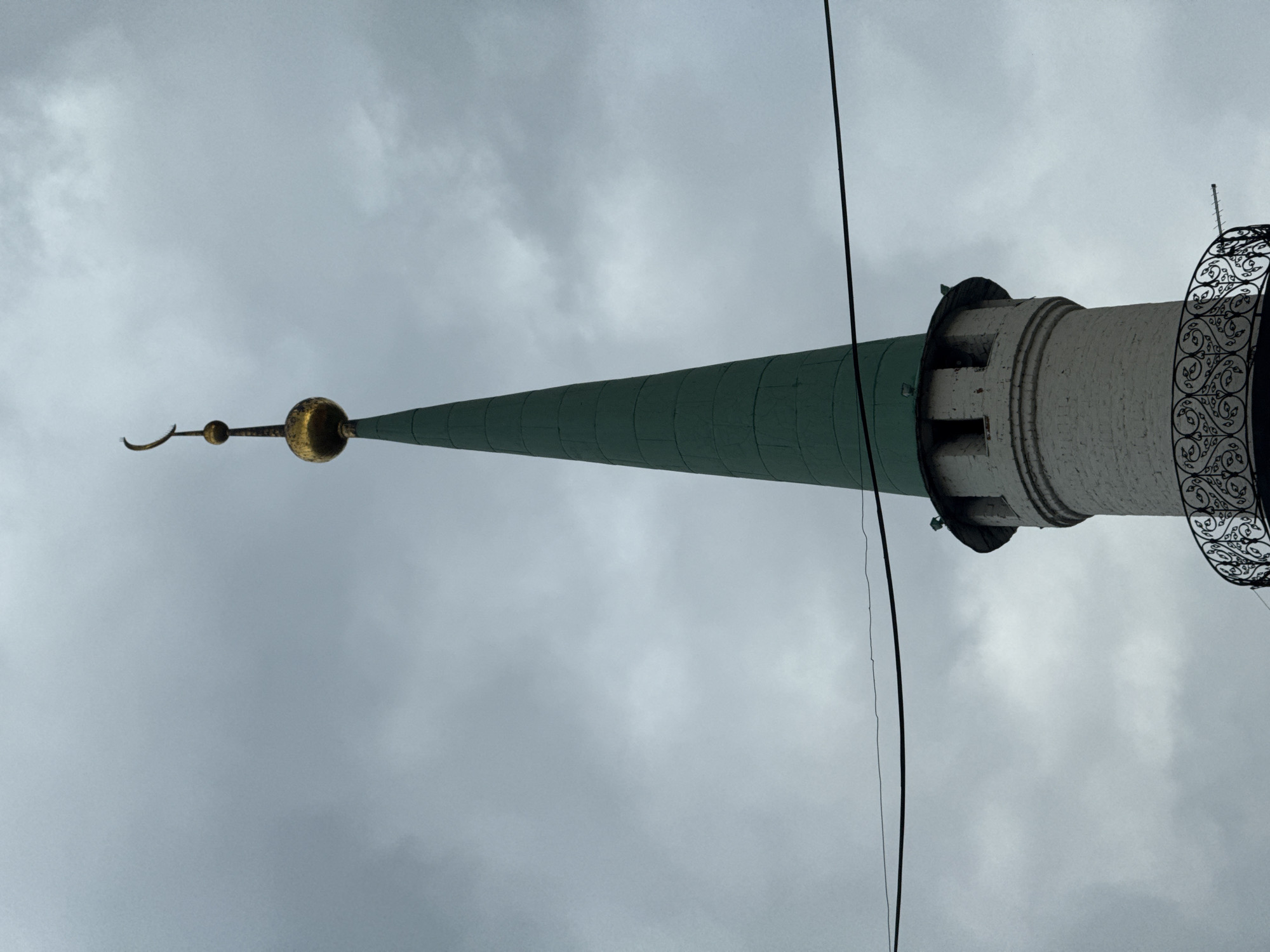 The image shows the top portion of the Kul Sharif Mosque minaret in Kazan, Russia, against a cloudy sky. The minaret is tall and slender, with a conical green roof topped by a gold-colored finial featuring a crescent moon. The lower part of the minaret is made of white brick and has a decorative black metal balcony. A black wire runs horizontally across the upper portion of the image. The overall impression is one of architectural grandeur under a muted sky.
