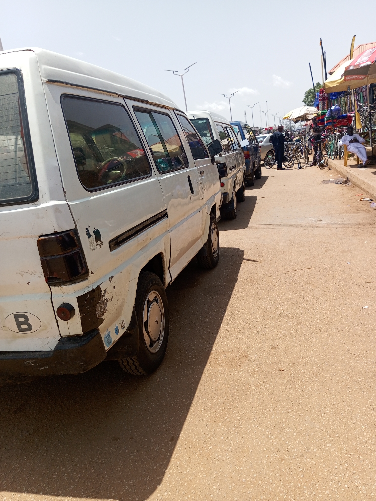 The image depicts a street scene in Bukuru, Nigeria, likely during the daytime under a clear sky. The foreground is dominated by the side of a white van, showing signs of wear and tear. It has a black bumper, a taillight assembly, and a circular sticker with the letter "B" on it. To the right of the van, a portion of the road surface is visible, appearing to be asphalt.
Further down the street, a line of parked vehicles, including more vans and cars, are visible. The scene then transitions to a marketplace area on the right side of the street. Numerous stalls are set up, displaying various goods such as bicycles, bags, and other merchandise. Several people are present, some interacting with vendors or browsing the items. One man, dressed in white traditional attire, is seated on a yellow stool, observing the surroundings. Another person, also in traditional clothing, appears to be looking at bicycles.
Notable details include the presence of multiple bicycles, suggesting it might be a hub for their sale or repair. The stalls are covered with umbrellas and awnings to provide shade. The overall atmosphere appears to be that of a busy market day. The time of day is indicated by the bright sunlight and the long shadows cast by the vehicles. There are no explicit indications of weather other than the clear sky. No visible text is discernible in the image apart from the "B" sticker on the van.