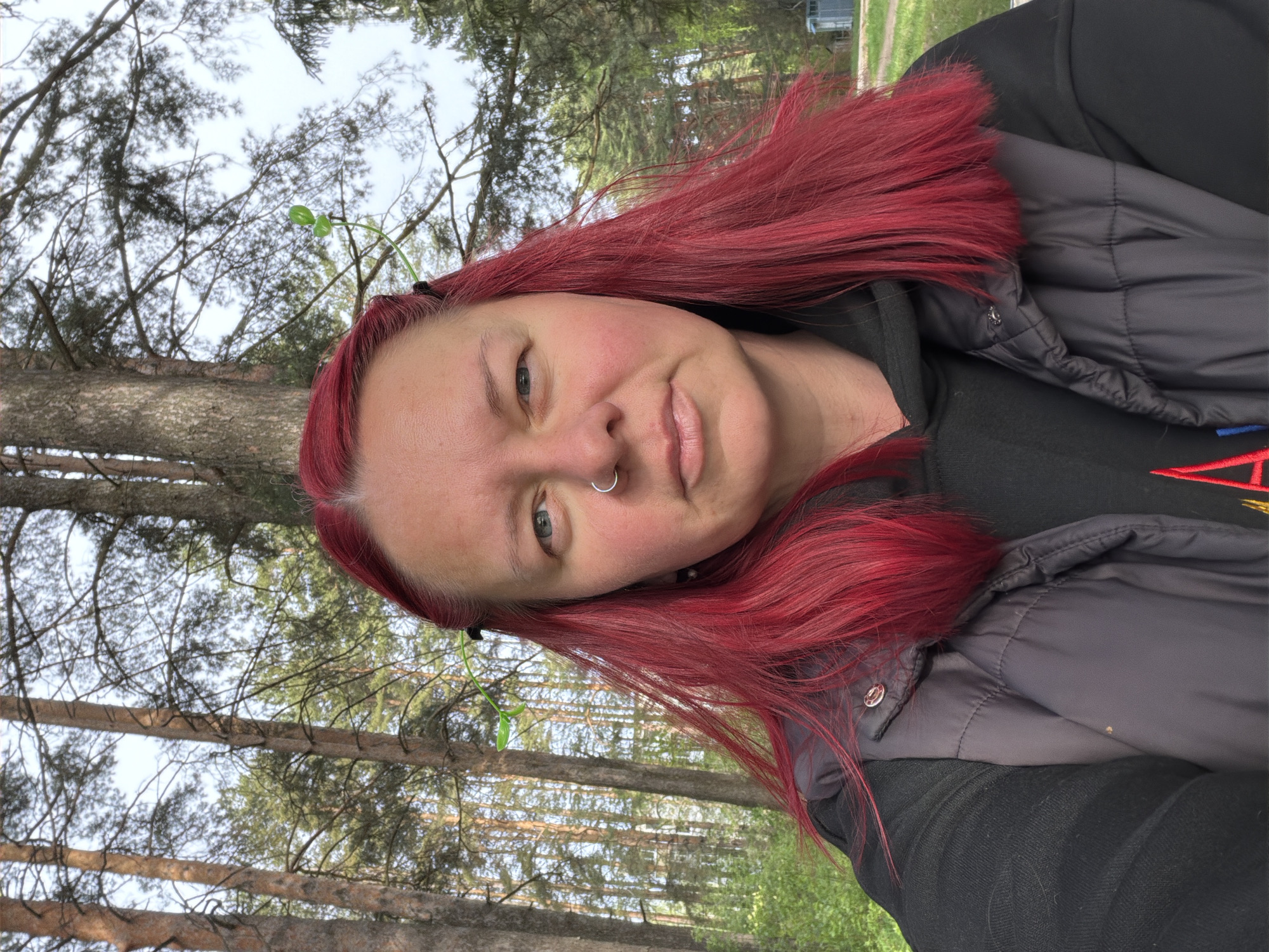 A woman with red hair, a nose ring, and wearing a black jacket is shown outdoors in a forested area. Tall trees with light brown trunks are visible in the background. One tree is close on the left. A small sprig with leaves is positioned on her head.