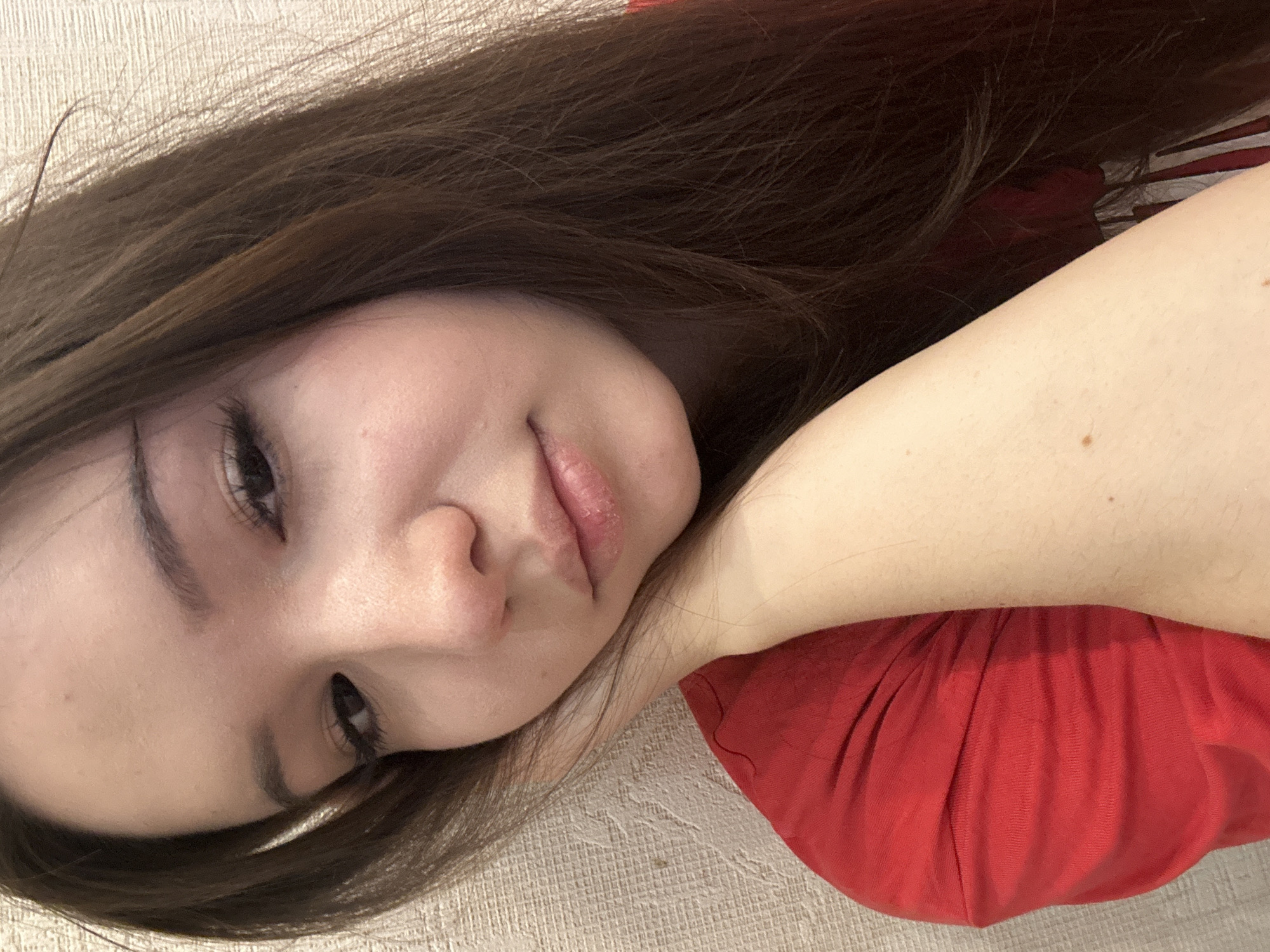 A young female is depicted in a close-up, head-and-shoulders portrait, positioned indoors in the city of Naberezhnye Chelny, Russia. She has long, dark brown hair, parted on the left side, with strands falling forward and obscuring part of her right shoulder and arm. Her head is tilted slightly to her left, resting on her bent left arm, with her left hand supporting her chin and lower jaw. Her eyes are dark, with visible long eyelashes, and are directed downwards and to her left. Her eyebrows are dark and well-defined. The skin on her face shows natural texture with minor imperfections visible on her forehead and nose. Her lips are slightly parted, revealing some chapping or dryness on the lower lip. She is wearing a red short-sleeved garment, likely a t-shirt, with fabric folds evident on her left shoulder and arm. A light-colored, textured wall serves as the background, exhibiting a fine, somewhat horizontal patterned surface. A small mole is visible on her right upper arm, which is partially visible on the bottom right of the frame.