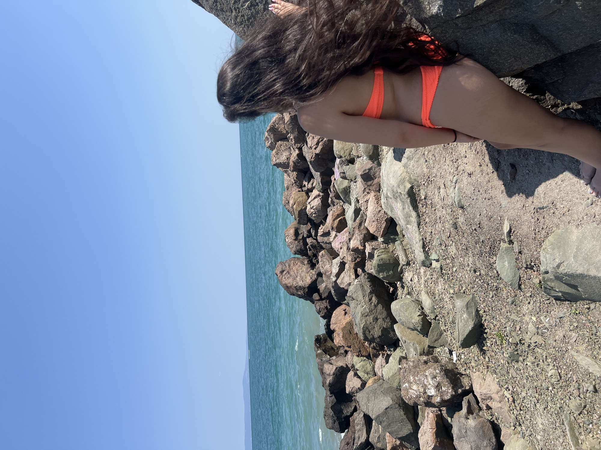 A person in an orange bikini stands on a rocky shore in Pomorie, Bulgaria, facing the sea. Their long, dark hair is blowing in the wind. The sea is a turquoise color, and there are large rocks and pebbles on the shore. The sky is clear and blue.