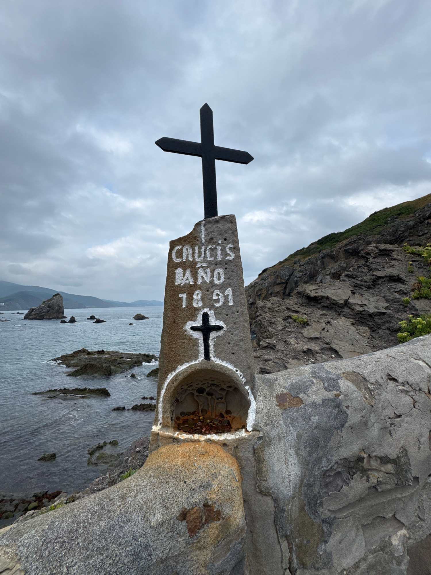 A stone cross memorial stands on the coast of Bermeo, Spain, under a cloudy sky. The memorial has a larger black cross at its apex, and below it, inscribed in white lettering, are the words "CRUCIS BAÑO" and the numerals "1891". A smaller black cross is painted below the inscription, and a small alcove at the base of the memorial contains a collection of coins. The sea is visible in the background with rocky outcrops. A steep, vegetated hillside rises behind the memorial. The memorial is built on a stone wall overlooking the water.