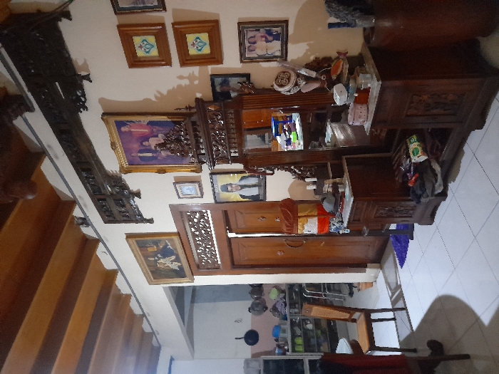 The image shows the interior of a home in Mojokerto, Indonesia, located at the coordinates -7.459 latitude and 112.447 longitude. The room appears to be a living room or bedroom with a large wooden dresser with an ornate mirror. The dresser is filled with various items, including toiletries and decorative items. The room has a white tiled floor. There is a wooden door to the left of the dresser. There are framed pictures on the wall behind the dresser. The walls are painted white and there is a wooden ceiling with beams. There are wooden shelves on the left side of the room and a large wooden table with a chair in the foreground.
