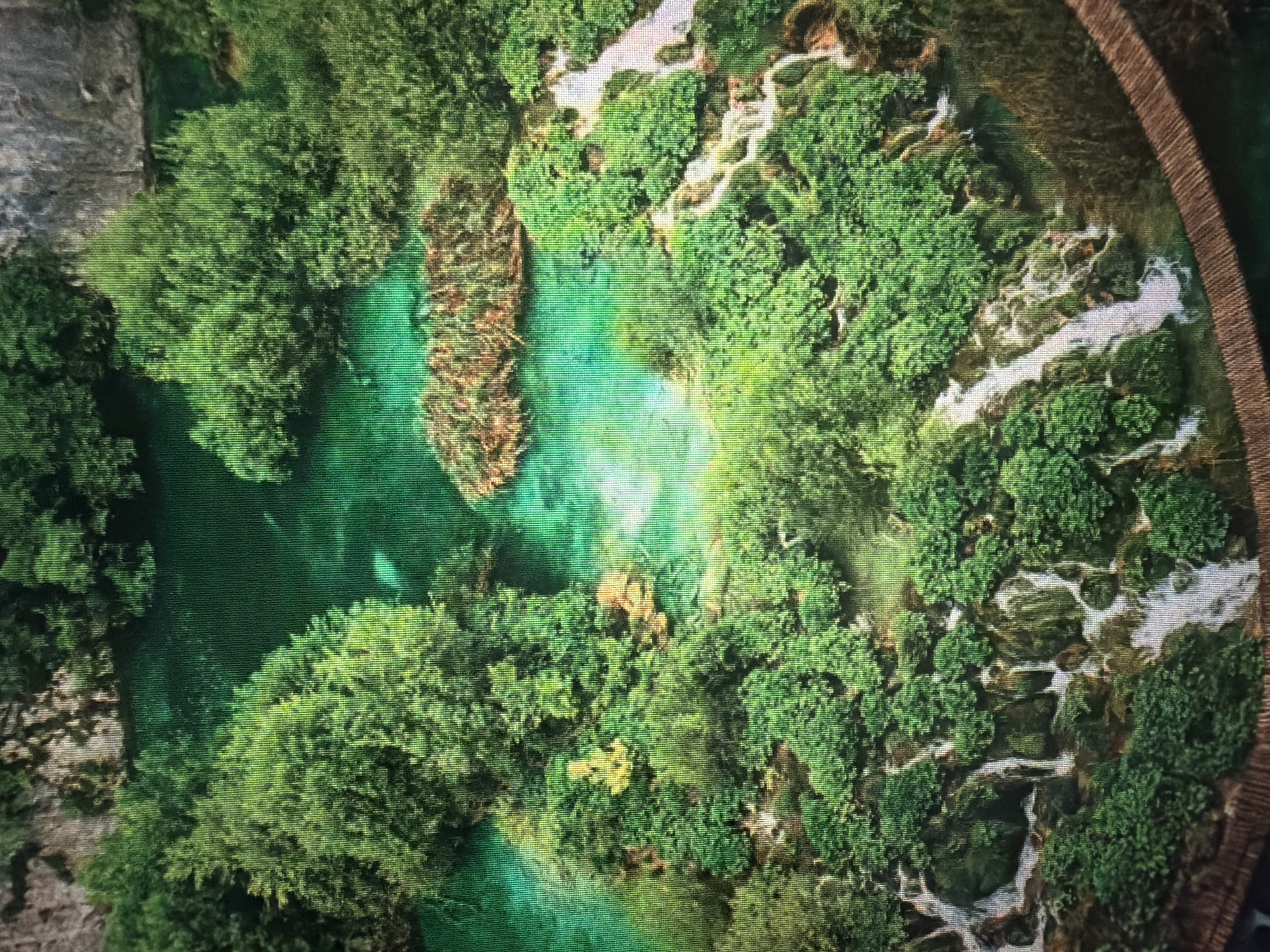 The image captures an aerial view of a vibrant, multi-tiered waterfall cascading into a clear, turquoise body of water. Lush green trees and dense foliage surround the water features, creating a serene natural landscape. The water appears shallow in some areas, revealing submerged rocks and sandy bottoms. In the lower right portion of the image, a wooden boardwalk or bridge is visible, hinting at a well-maintained tourist path. The overall scene is one of unspoiled natural beauty. The location is identified as Baan Mae Nam, Thailand.
