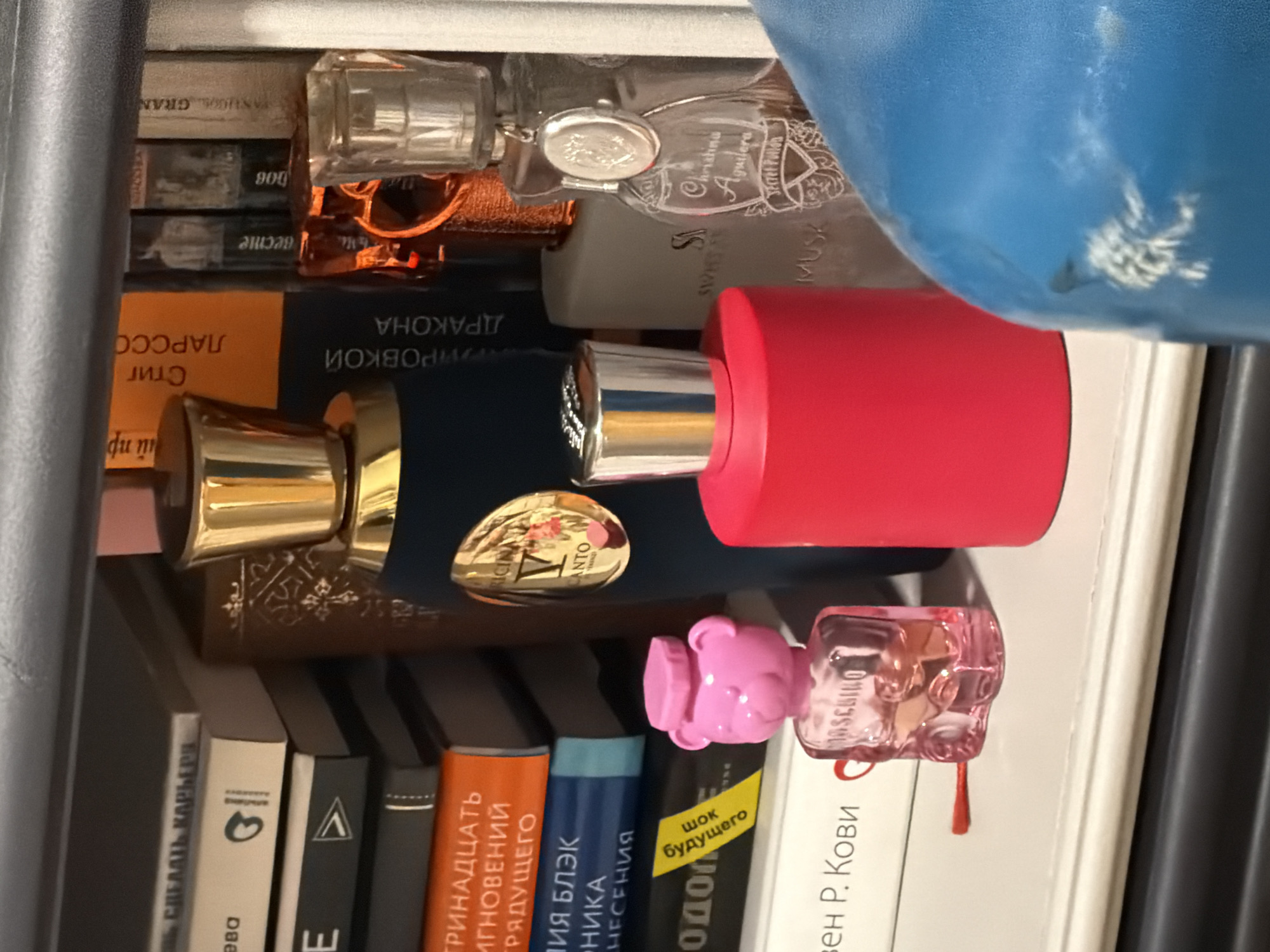 The image shows a shelf stacked with books and various perfume bottles. The books are mostly in Russian, with titles such as "СДЕЛАТЬ КАРЬЕРЯ," "ТРИНАДЦАТЬ ИГНОВЕНИЙ," "ДРАКОНА," and "ЛАРССО." Several perfume bottles are visible, including brands such as Christina Aguilera, Moschino, and a bottle with "V Canto" labeled on it. The bottles vary in shape and color, some being clear, others pink or red, and one with a gold-colored top. A corner of a blue object is visible in the upper right. The setting appears to be a private shelf, possibly in a home. There is no clear indication of Moscow or Russia other than the Russian language on the book titles.