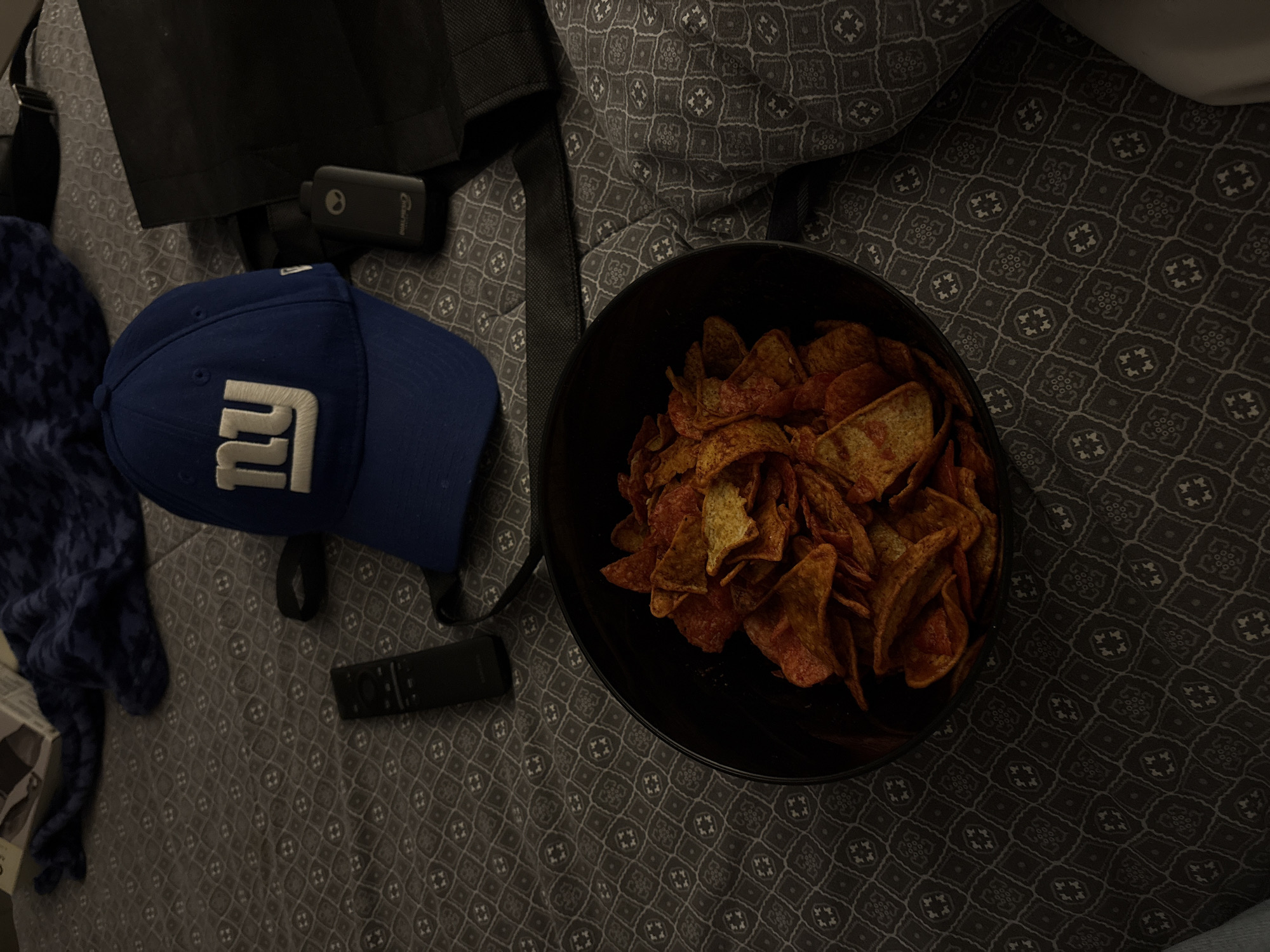 The image shows a dimly lit room with a bed covered in a patterned grey and white bedspread. A black bowl filled with reddish-brown tortilla chips sits on the bed. Adjacent to the bowl are a blue baseball cap with the letter "NY" on it, a black bag with straps, a small black electronic device, and a black remote control. A blue patterned scarf is visible on the left side of the image.
The location is not discernible from the image, and there is no information to confirm that the image was taken in San Bernardino Tlaxcalancingo, Mexico.