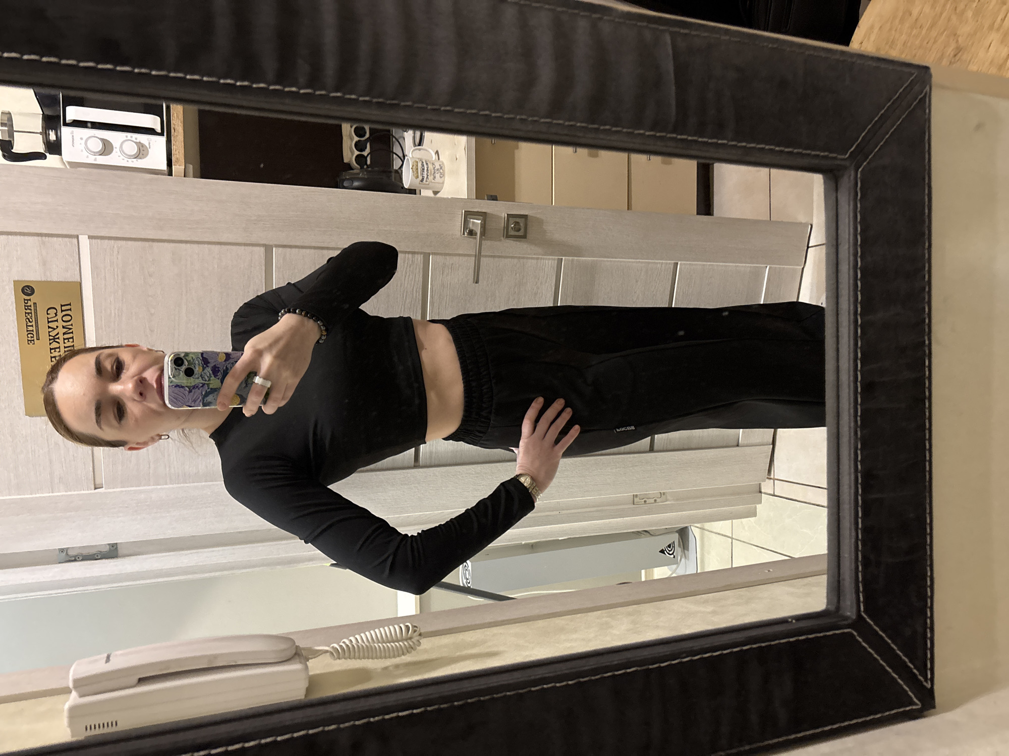 A reflected view through a dark-framed mirror shows a woman taking a selfie in what appears to be an apartment or hotel room of Unknown, Unknown. The woman is dressed in a black long-sleeved crop top and matching black pants. She is holding her phone up with her left hand, and her right hand is resting on her hip. A landline phone is mounted on the wall and is visible near the bottom left corner of the mirror. Behind her in the reflection is a door with a silver handle and lock set. Above the door, there is a white microwave and a mug on a shelf. The wall color is a neutral beige.