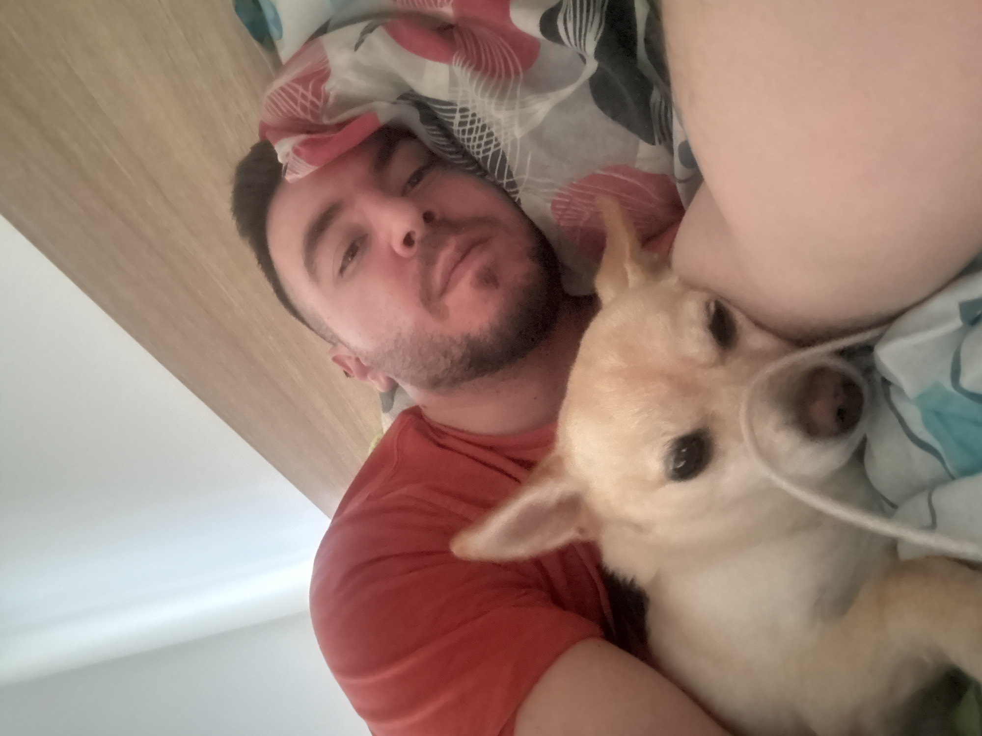 The image depicts an indoor scene within a private residence in Sofia, Bulgaria. An adult male is lying on his left side in a bed, facing the camera. He has short dark hair, dark stubble, and wears a red t-shirt. His head is partially covered by a patterned duvet featuring red and black circular designs on a white background. Adjacent to him, a small, light-tan colored dog with erect ears is also lying down, resting its head on the man's arm or body. Its eyes are partially closed. A white cord lies across the dog's muzzle. The man's left arm and a light blue and white striped sheet are visible below the dog. A light wooden headboard is visible behind the man's head, and a white ceiling is partially visible in the upper left background. The perspective indicates a self-portrait (selfie).