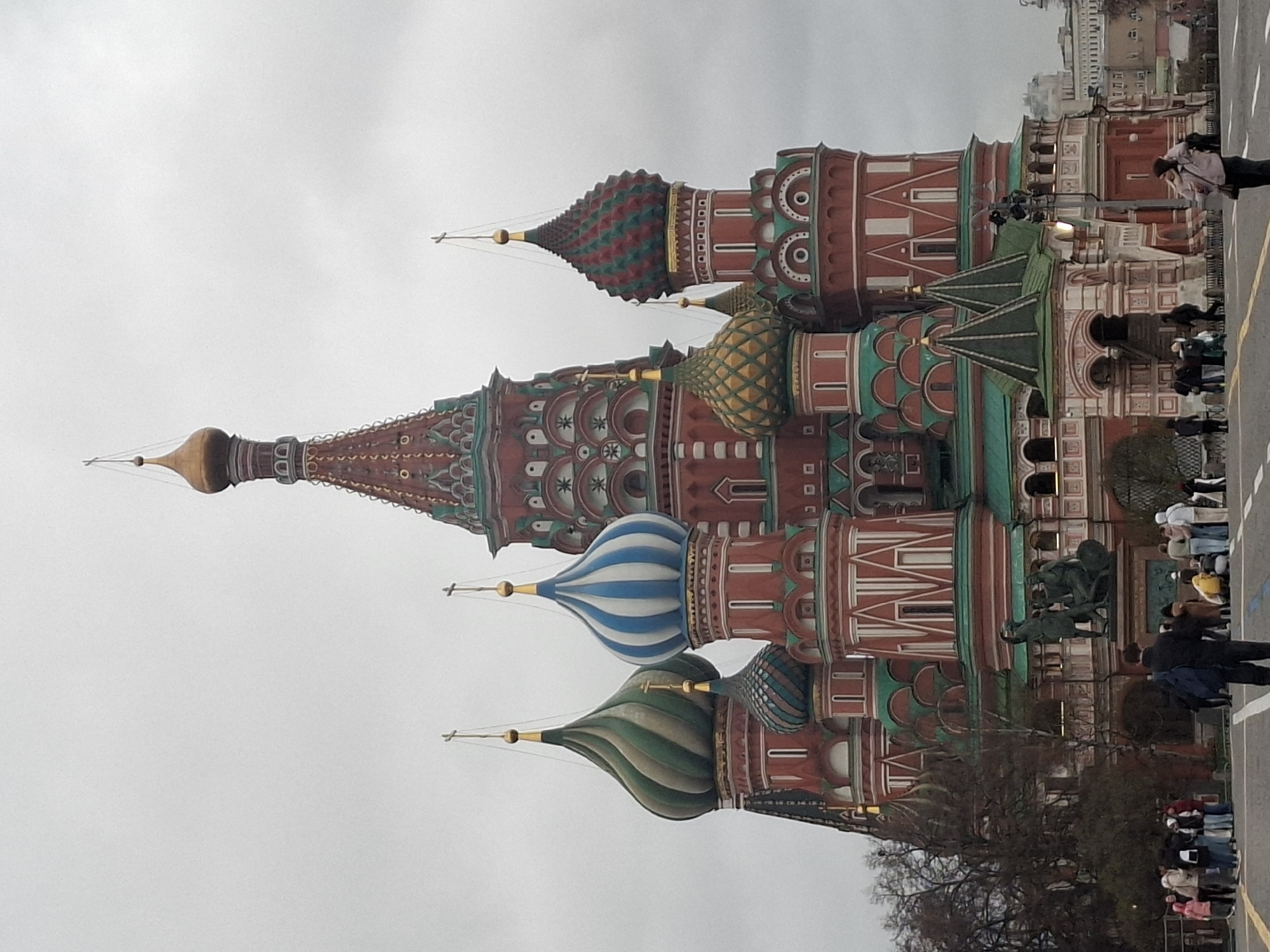 The image shows the iconic Saint Basil's Cathedral in Moscow, Russia. The cathedral's colorful onion domes are prominently displayed, against a gray sky. Pedestrians are walking along a sidewalk in front of the building. A bronze monument is also visible.