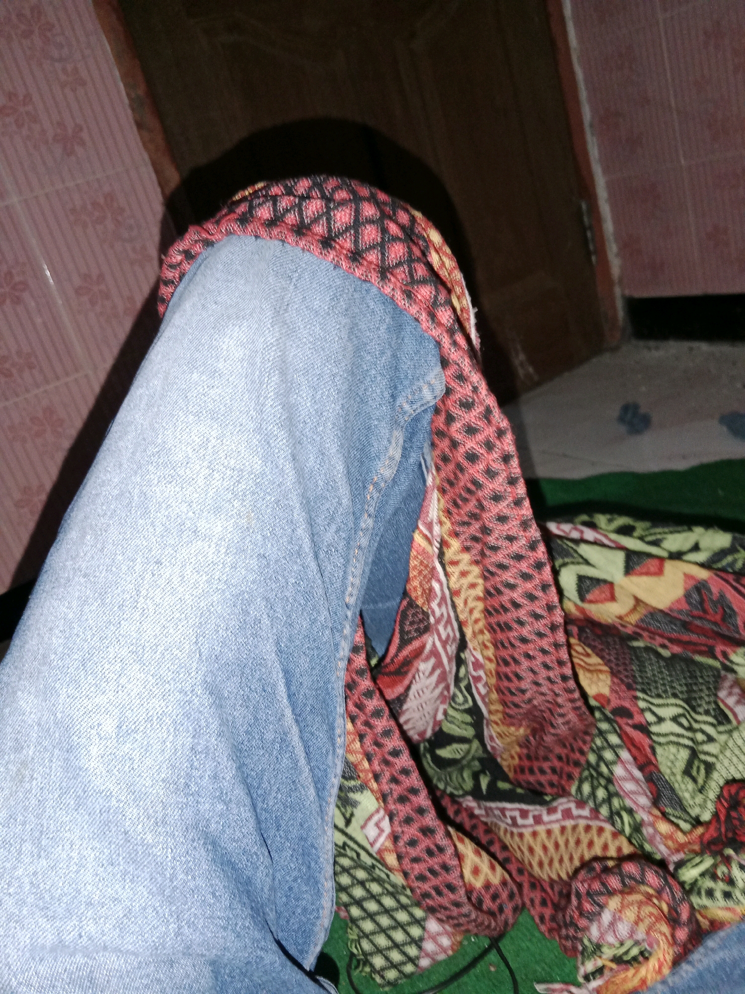 The image shows a close-up view of a person's left leg, clad in blue denim jeans. A brightly colored patterned fabric, featuring red, black, yellow, and green geometric designs, is draped over the leg, partially obscuring it. The fabric appears to be a scarf or a blanket. The background is indistinct, with a wall on the left that has a pink, patterned texture and a darker, possibly wooden surface on the right. The scene suggests an indoor setting, possibly a bedroom or living area. There is no visual information to confirm the location in Ringinrejo, Indonesia.