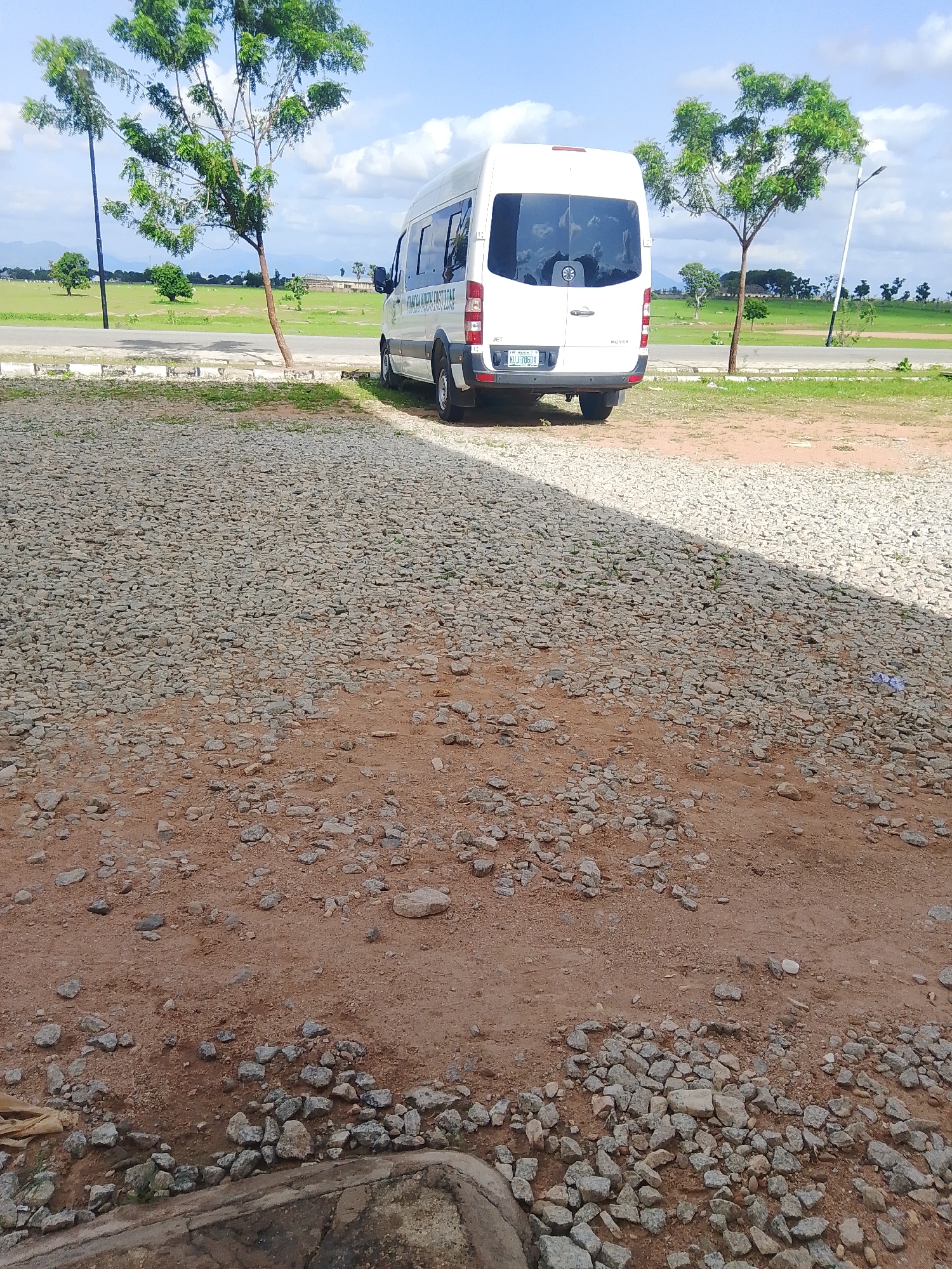 A white Mercedes-Benz Sprinter van is parked on a gravelly lot, facing away from the camera. The van has tinted windows and a license plate that reads "KUJ 706GX". On the side of the van, text is visible, likely indicating a company name or service. The van is parked in a somewhat overgrown area with patches of grass and dirt.
In the background, there are several trees with green foliage, some appearing larger and more mature than others. Beyond the trees, a vast, open grassy field stretches out, with some distant buildings and rolling hills under a bright, partly cloudy sky. A paved road runs along the edge of the gravel lot and the field. A lamppost stands tall on the right side of the frame. The overall scene suggests an outdoor setting, possibly a rural or semi-urban area, during daylight hours. The lighting indicates it is likely daytime. The terrain is rough and uneven, with a mix of gravel, dirt, and sparse vegetation.