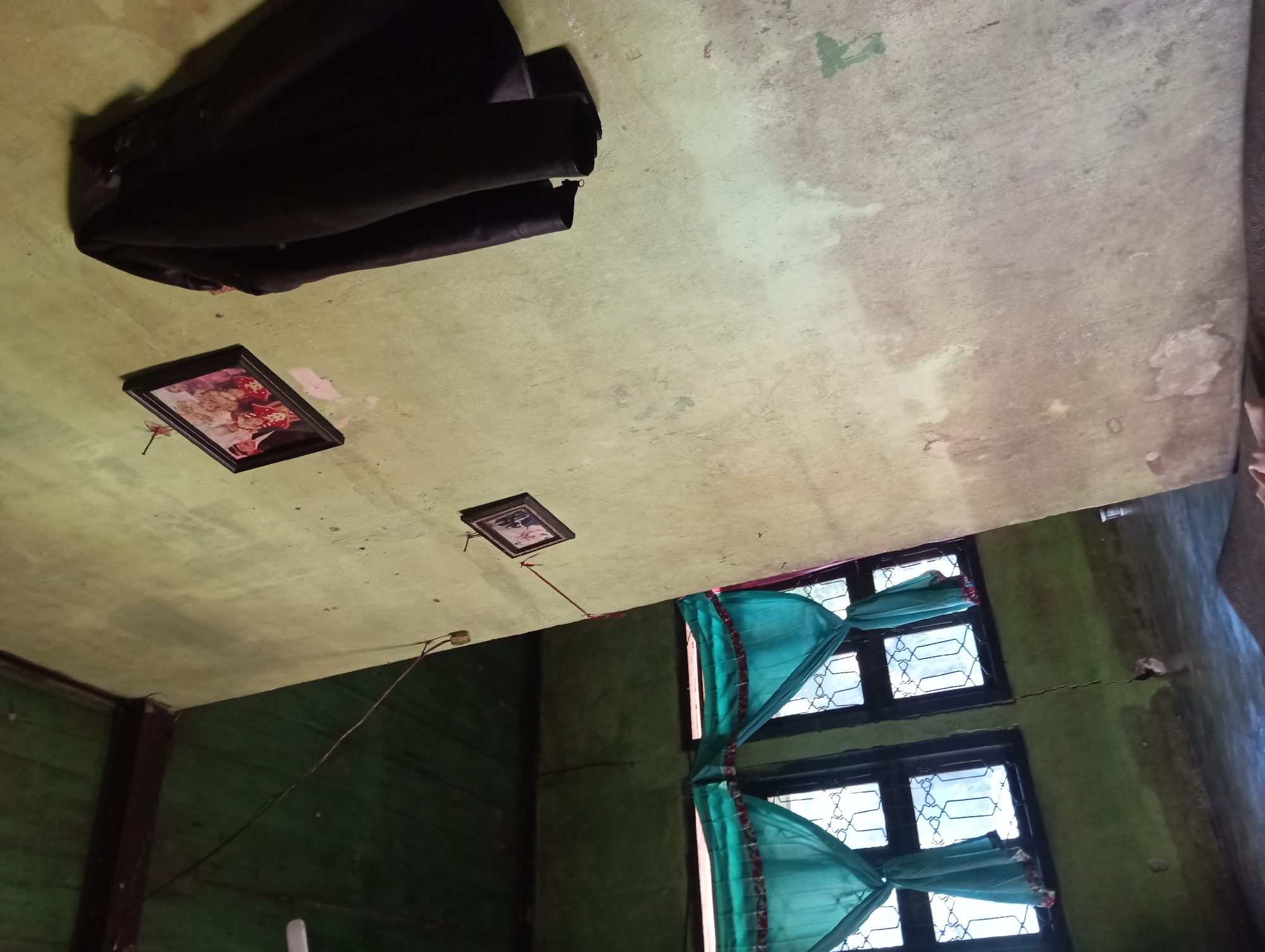 The image depicts an interior scene of a room in Rejosari, Indonesia. The room appears somewhat aged, with green painted walls and a dark green ceiling. A black jacket is draped over the wall on the right side of the frame. Two framed photographs hang on the wall: one larger, at the top, and a smaller one below it. In the background, on the left, two windows are visible, each covered with teal-colored curtains that have a patterned trim. The windows have dark frames and ornate metalwork on the glass panes. The floor is dark and reflective, suggesting it might be wet or polished. A crack is visible in the wall near the windows. The lighting suggests it is daytime, but the overall atmosphere is subdued. There are no people visibly present in the scene.