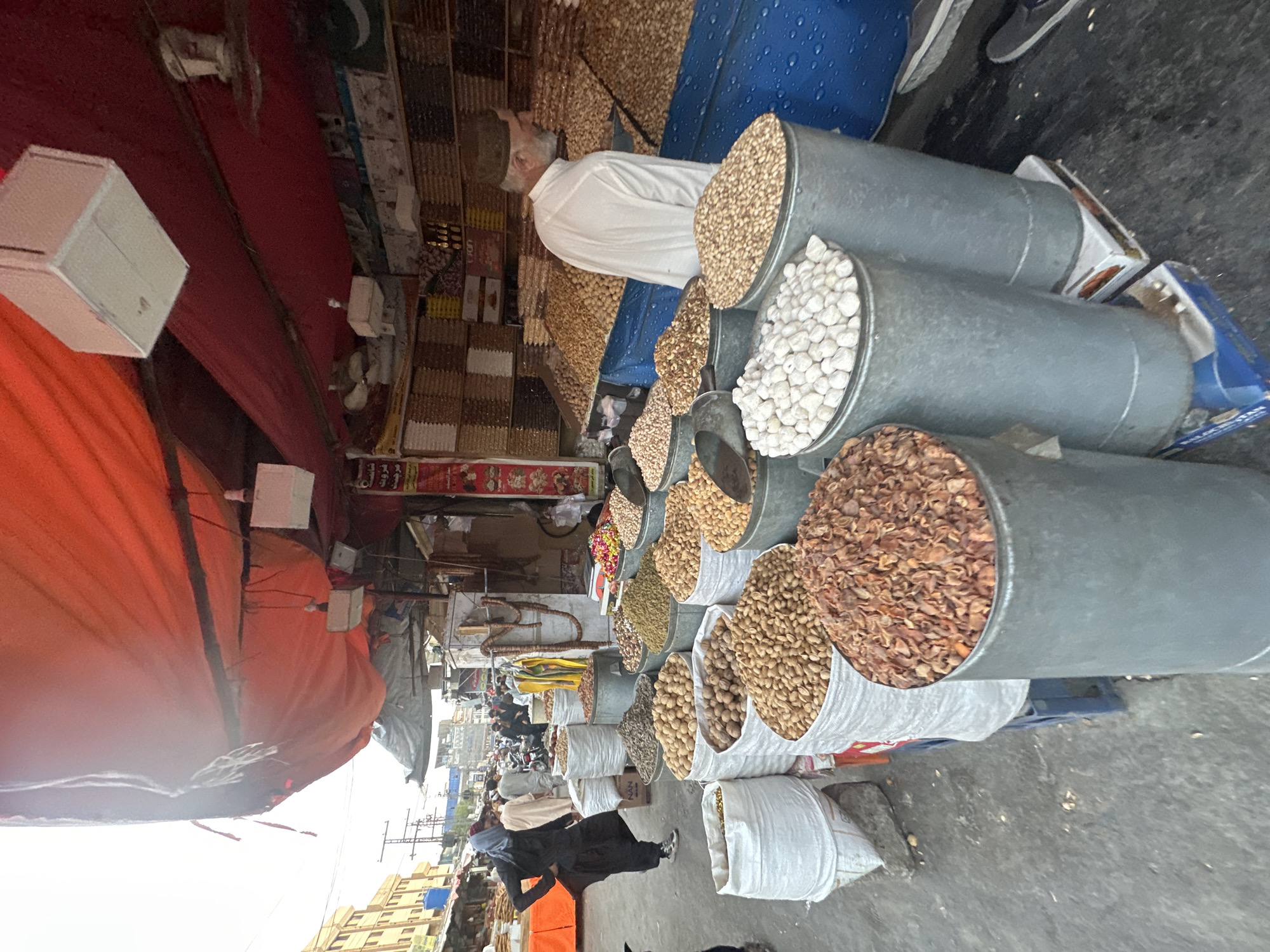 The image shows a street market in Quetta City Tehsil, Pakistan. A vendor stands behind a display of nuts and dried goods, arranged in metal containers and bags. He is wearing a white shirt and a hat. There are shelves filled with various products behind him. An orange awning covers part of the market. People are walking past the stall. Buildings are visible in the background. The sky is overcast.
