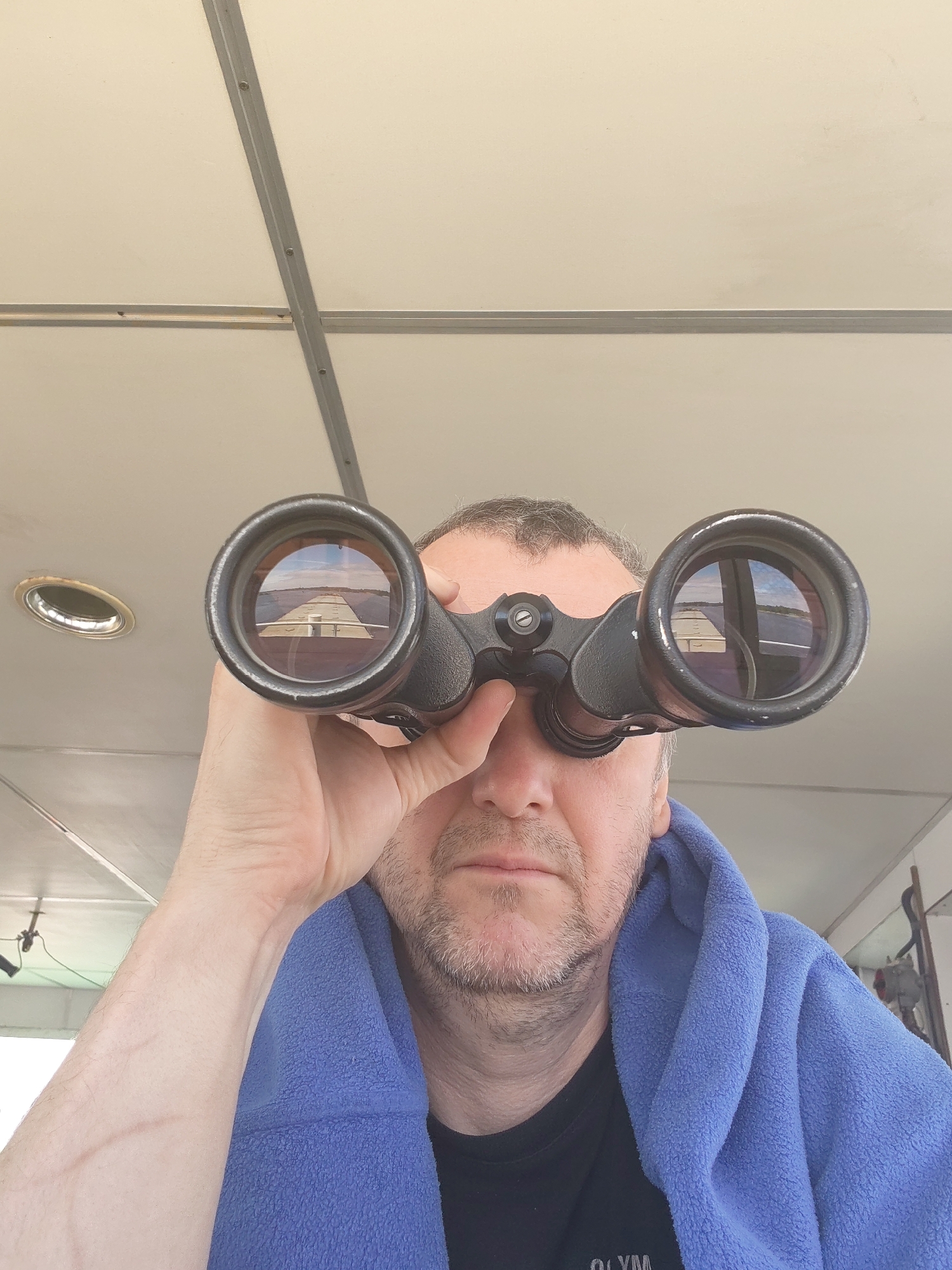A middle-aged man is looking through a pair of black binoculars. The reflection in the left lens shows a two-story building with a grey roof and white walls, while the reflection in the right lens shows trees and sky. The man is wearing a blue terrycloth robe and a black t-shirt with white writing. The background shows a white ceiling with metal crossbeams. The location is likely Kargulino, Russia, based on the reflection in the binoculars.