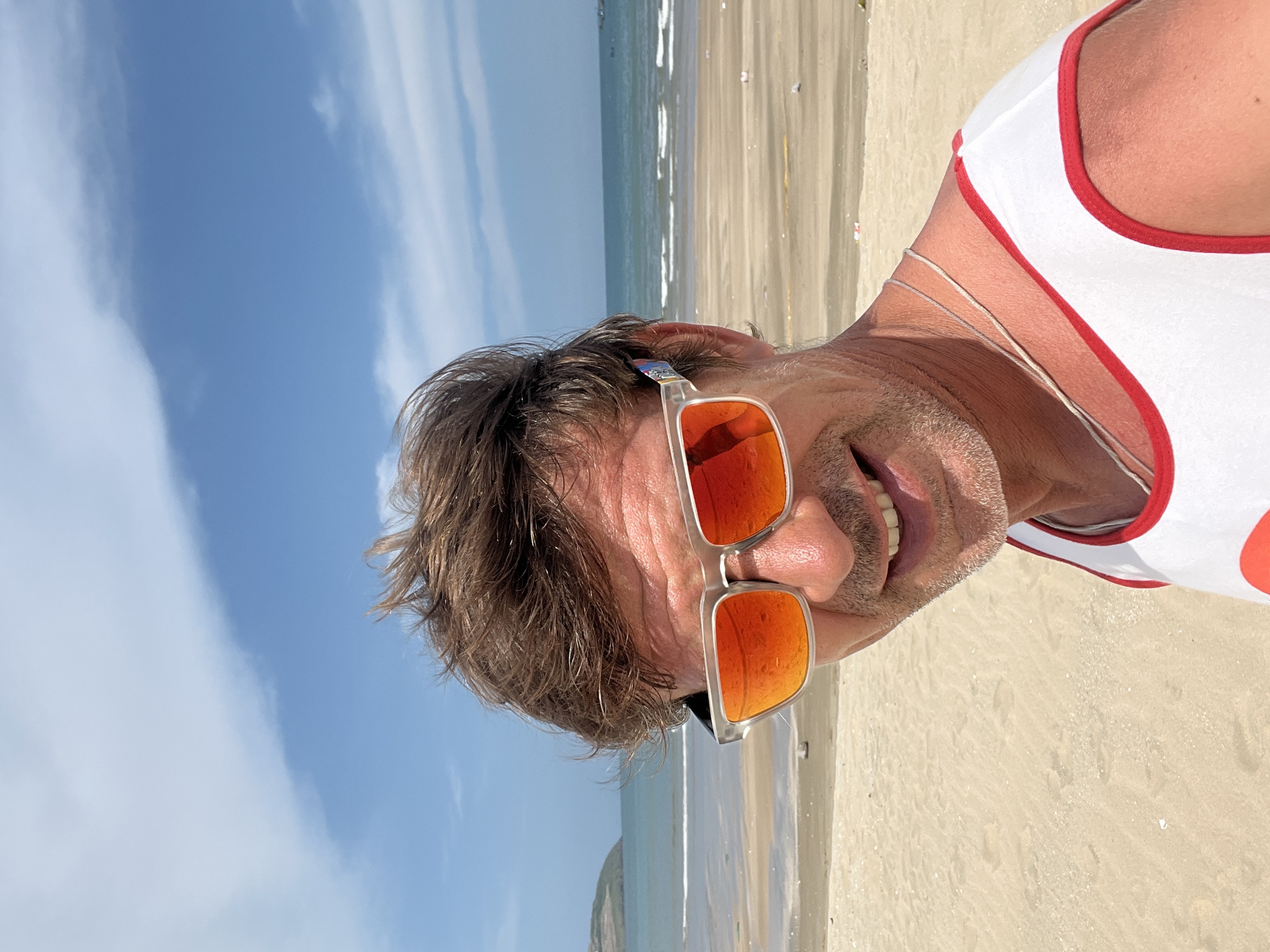 A man wearing sunglasses and a white tank top with red trim stands on a sandy beach in Mui Ne, Vietnam. The sky is blue with scattered white clouds. The ocean is visible in the background with gentle waves. The sand is light brown and appears wet in some areas. Some debris is scattered on the beach. The man is smiling and looking at the camera. His hair is brown and tousled, and he has a light beard. He is wearing a thin silver chain around his neck.
