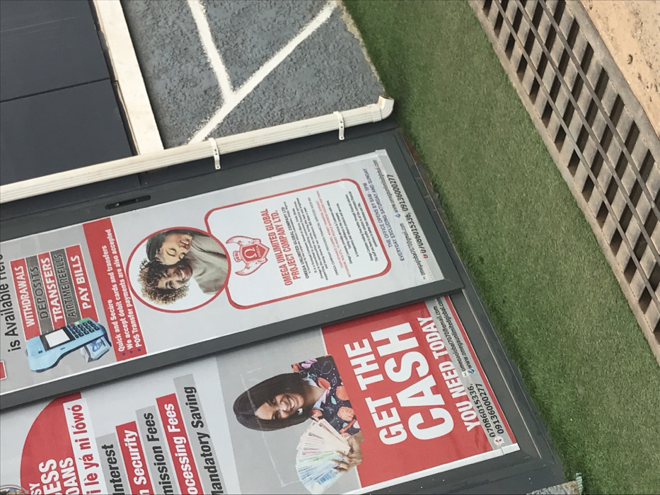 The image displays two vertical advertising banners affixed to the exterior of a commercial building in Akure, Nigeria. The banners promote "OMEGA UNLIMITED GLOBAL PROJECT COMPANY LTD." The left banner, predominantly red and white, features a smiling woman holding a fan of Nigerian Naira banknotes with the prominent text "GET THE CASH YOU NEED TODAY." Partial text mentions "ESS LOANS," "Interest," "Security," "Mission Fees," "Processing Fees," and "Mandatory Saving." The right banner, white with red accents, lists services such as "WITHDRAWALS, DEPOSITS, TRANSFERS, AIRTIME REFILLS, PAY BILLS," accompanied by an image of a mobile POS terminal. This banner also includes a circular photograph of two smiling women and the company logo. Text below indicates "Quick and Secure," accepting "debit cards and transfers," and "POS Transfer payments." The company is identified as "OMEGA UNLIMITED GLOBAL PROJECT COMPANY LTD." based in "Akure, Ondo State, Nigeria." Office hours are stated as "9AM - 5PM EVERYDAY EXCLUDING SATURDAY AND SUNDAY." Both banners provide contact phone numbers (07086015336, 09136000277, U/0806015336), an email (omega.global7070@gmail.com), and a website (www.omegaunlimitedglobal.com). The building's exterior features dark gray paneling and a white vertical downspout. Below the banners, a strip of artificial green turf is visible, adjacent to a ground-level metal drainage grate in the foreground.