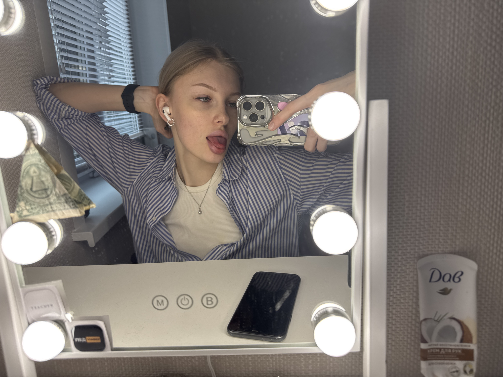 A selfie taken in a bathroom mirror. A young woman with blonde hair, wearing a blue and white striped shirt, is sticking her tongue out at the camera. She is holding a cell phone in her right hand to take the photo. The phone has a patterned case. The mirror is surrounded by lights. On the vanity below, there is a black smartphone, a small container, and a tube of Dove hand cream labeled with Cyrillic text, indicating the location as possibly Russia. There is also a folded US one dollar bill on the left side. The blinds are visible in the background. The city of Perm, Russia is not definitively discernible from the available evidence.
