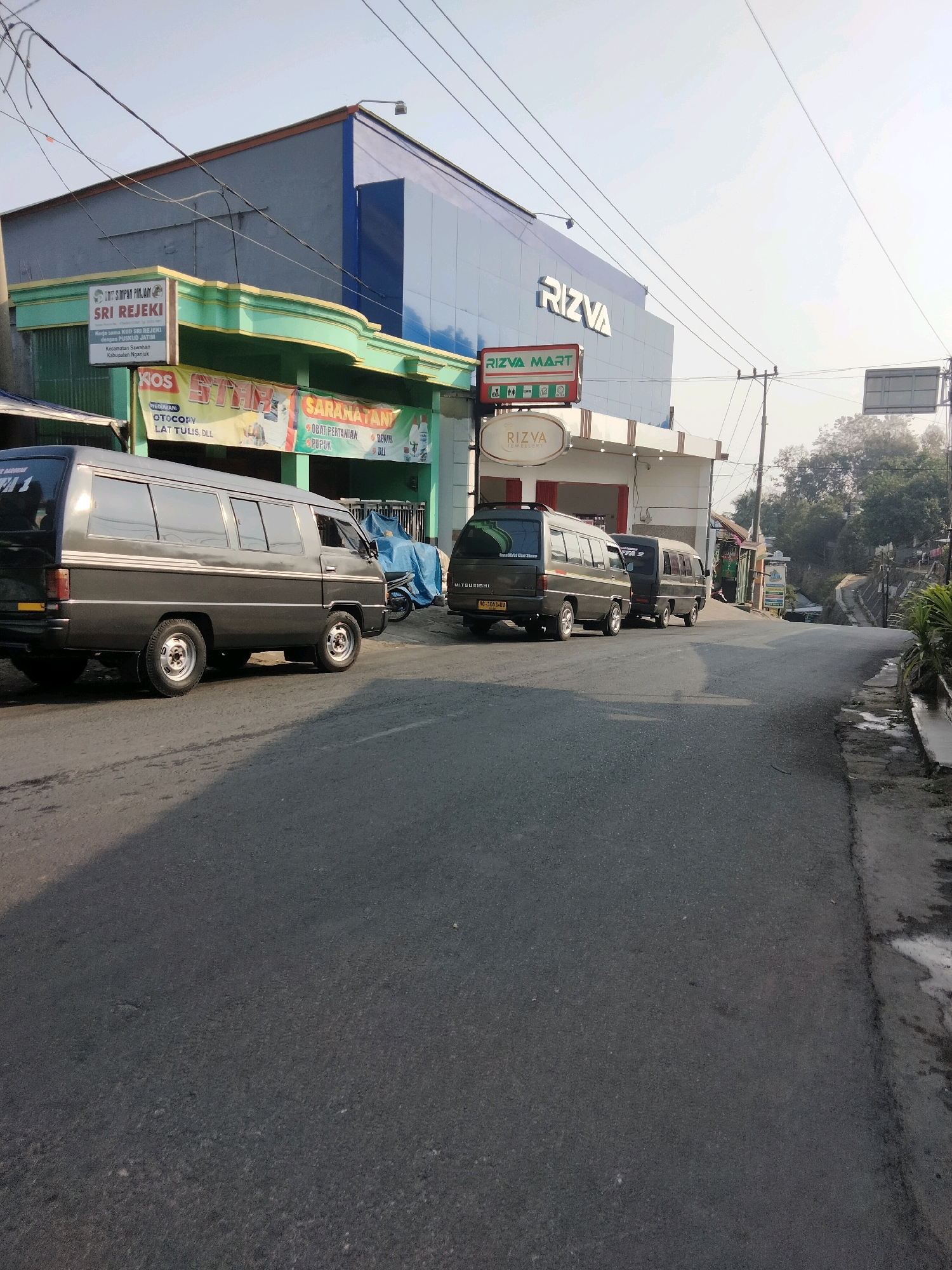 In the city of Sawahan, Indonesia, a paved road curves uphill. On the left side of the road, a building complex features a blue facade with "RIZVA MART" signage. Below this, a green awning shades a storefront with "SRI REJEKI" visible on a sign. Further down the awning, banners display services like "KOS," "KOTAK," and "SARANATAN." Several dark gray vans are parked on the right side of the road. The first van, a Mitsubishi, has its rear facing the camera. Two other similar vans are parked behind it. A blue tarp covers a motorcycle near the vans. Overhead, power lines crisscross the sky, which is bright and hazy. Trees and vegetation are visible on the uphill side of the road in the background. The overall scene depicts a commercial area with shops and transportation.