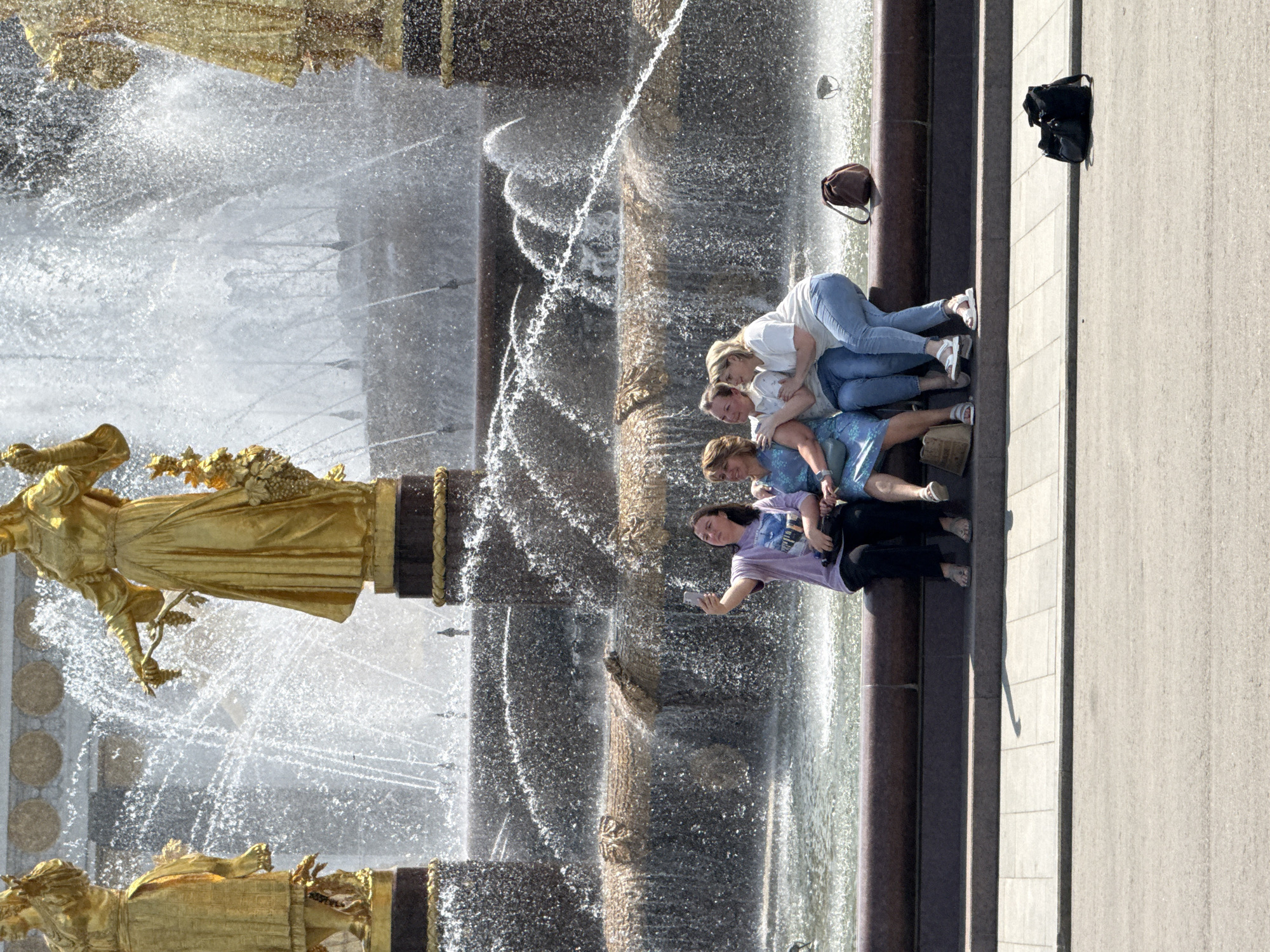 The image shows a fountain with golden statues in Moscow, Russia. Four women are positioned along the side of the fountain. The woman closest to the viewer is taking a selfie with her phone. The next woman is in a blue dress, followed by two women wearing white shirts and jeans, who are hugging. A dark bag is on the ground, and a brown bag is on the fountain. The fountain water is spraying upwards.