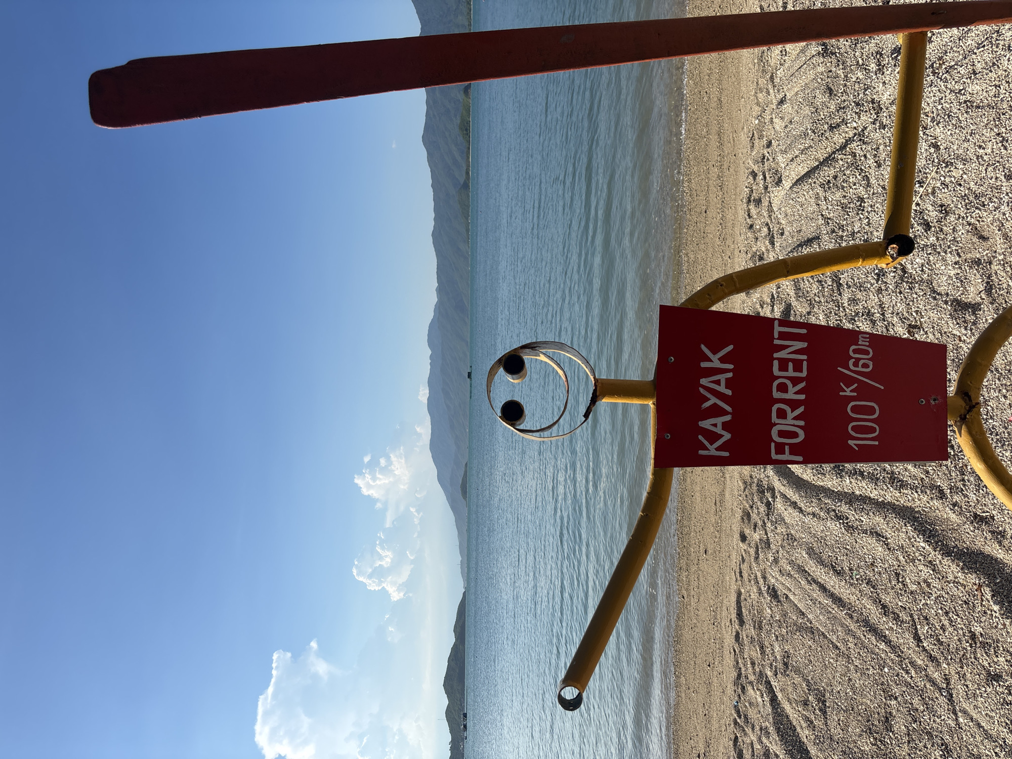 An outdoor shot captures a whimsical, homemade signpost on a beach in Vĩnh Lương, Vietnam. The signpost, fashioned to resemble a stick figure, is bright yellow with a red rectangular sign attached to its torso. The sign reads "KAYAK FOR RENT 100K/60m" in white lettering. The stick figure has a round, metal face with two large eyes and a smiling mouth. Its arms are outstretched, with one arm ending in a circular opening. To the right of the stick figure, a large, red kayak paddle stands upright, partially obscuring the background.
The beach itself is covered in a mixture of sand and small pebbles, with distinct patterns of light and shadow suggesting the late afternoon sun. Beyond the beach, the calm, blue ocean stretches out, meeting a line of verdant, tree-covered hills that form the horizon. The sky is a clear blue, dotted with a few fluffy white clouds. The overall atmosphere is one of a relaxed, seaside destination, likely a tourist spot offering recreational activities.