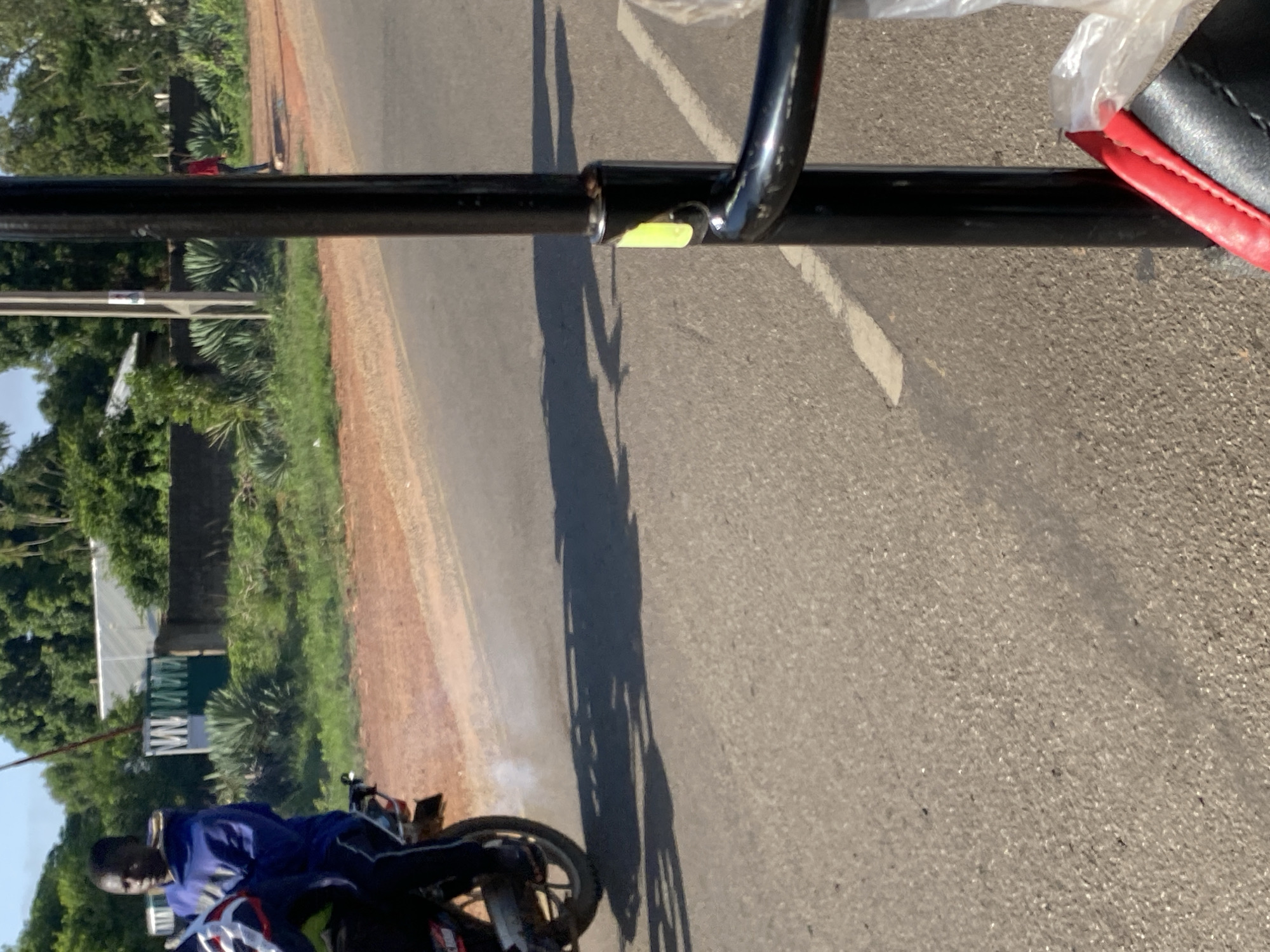 A young Black male wearing a blue hoodie and dark pants rides a motorcycle, emitting a puff of white exhaust. The motorcycle is partially obscured, but a wheel and part of the exhaust system are visible. The rider is in motion on an asphalt road, casting a shadow. The background shows a dirt shoulder with sparse vegetation and a low wall, beyond which are buildings and lush green trees under a clear blue sky. A black vertical pole, likely part of a vehicle or structure, occupies the right side of the frame. The scene appears to be in a rural or semi-urban setting in Nigeria.